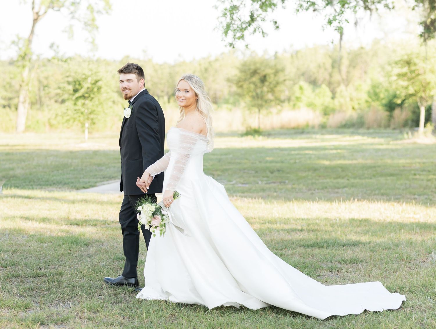 Bride in off-shoulder gown and groom in black tuxedo posing in sunlit green field on wedding day.
