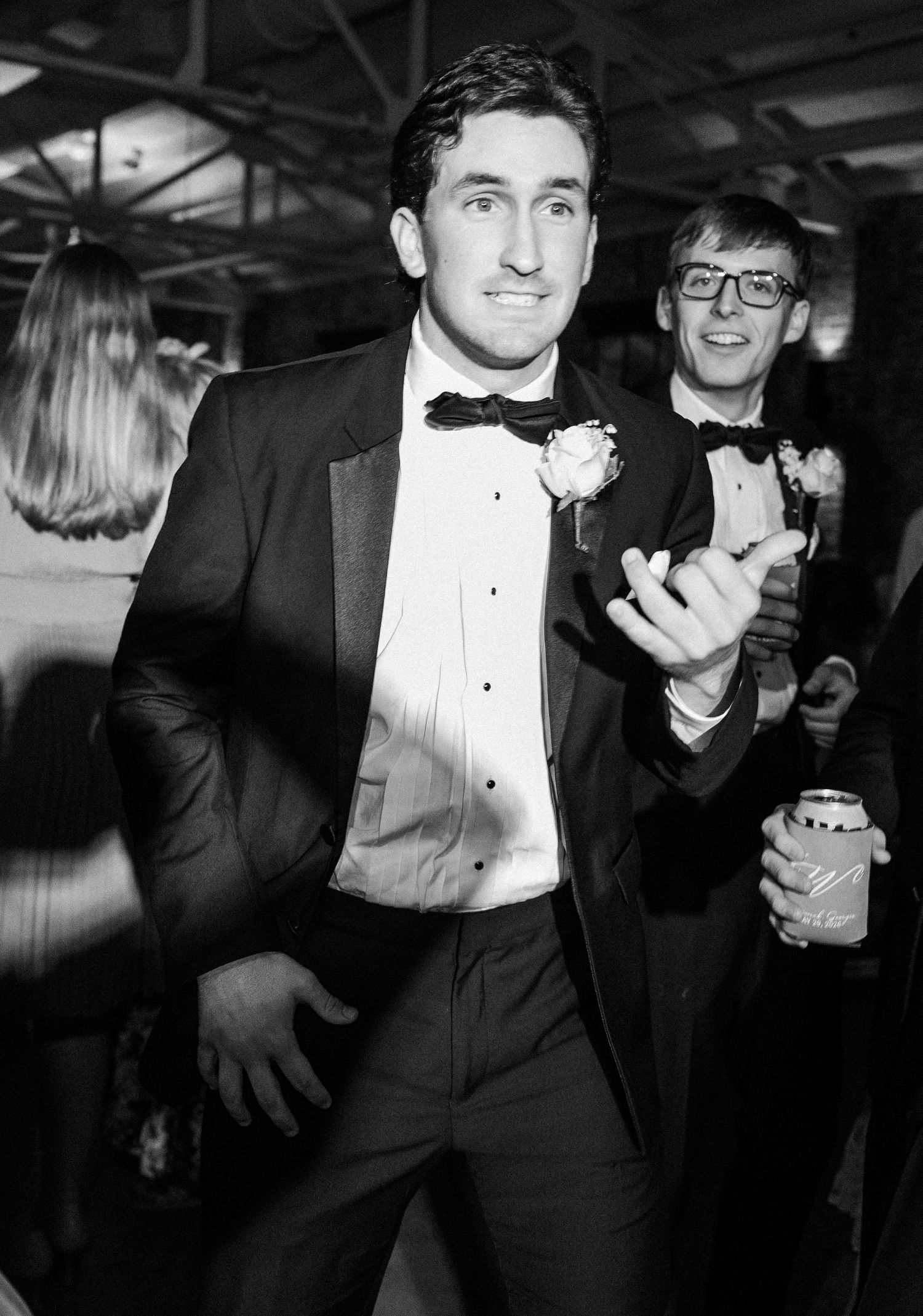 Groom in black tuxedo dancing energetically at wedding reception in black and white photo.