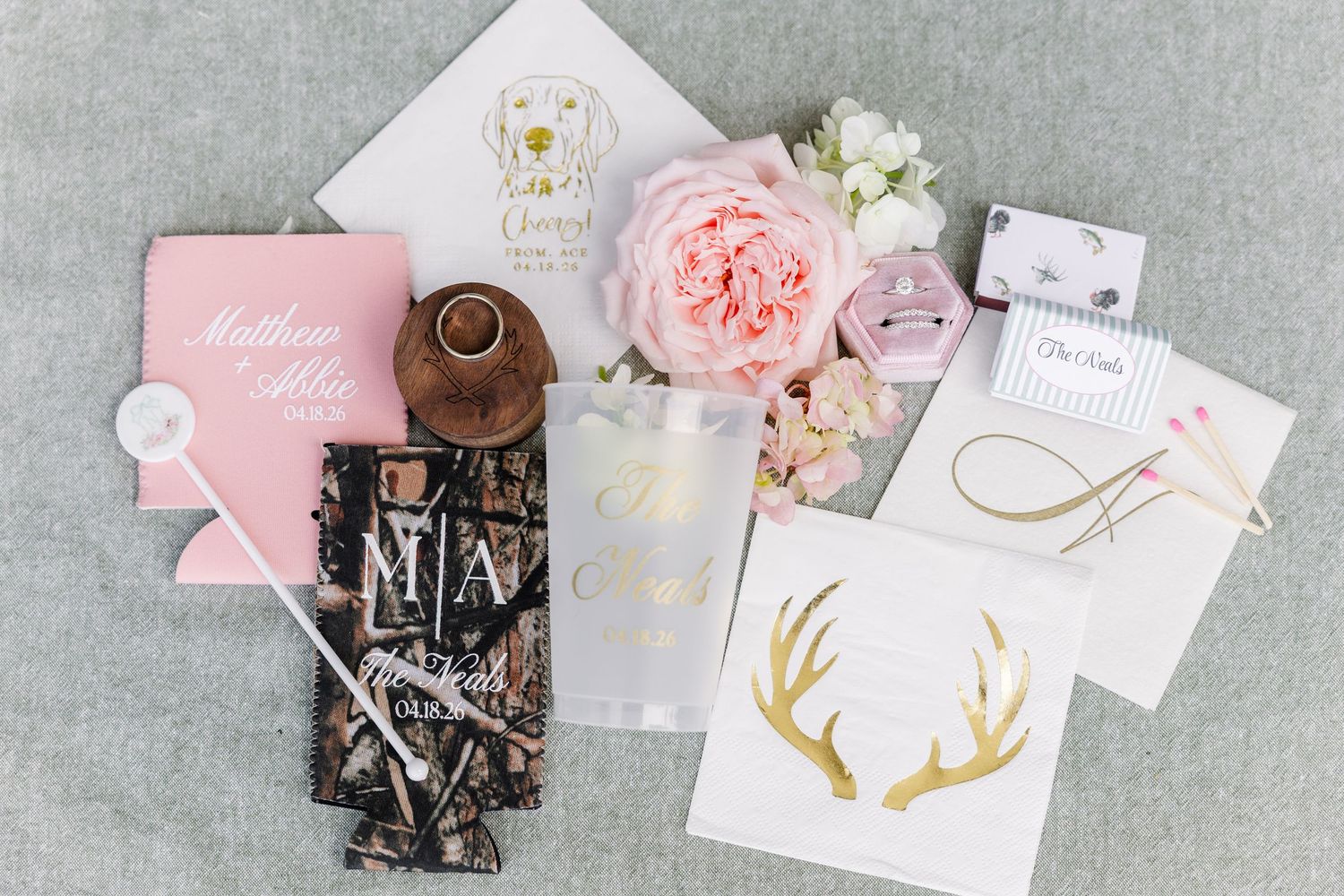 Pink and gold wedding flat lay with personalized gifts, floral accents, monogrammed items, and elegant stationery on gray background.