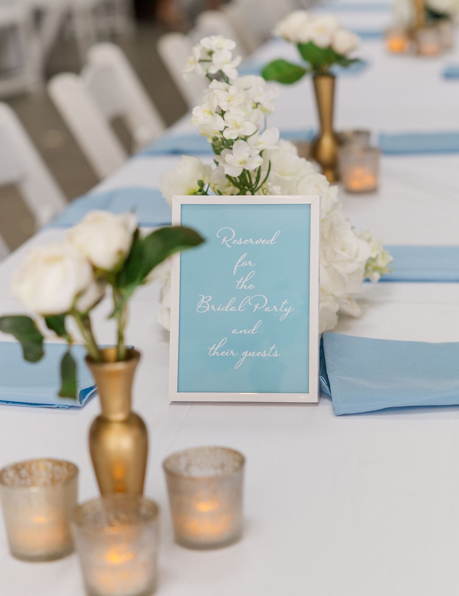 Elegant wedding reception table with blue napkins, gold vases, white roses, and a bridal party reserved sign.