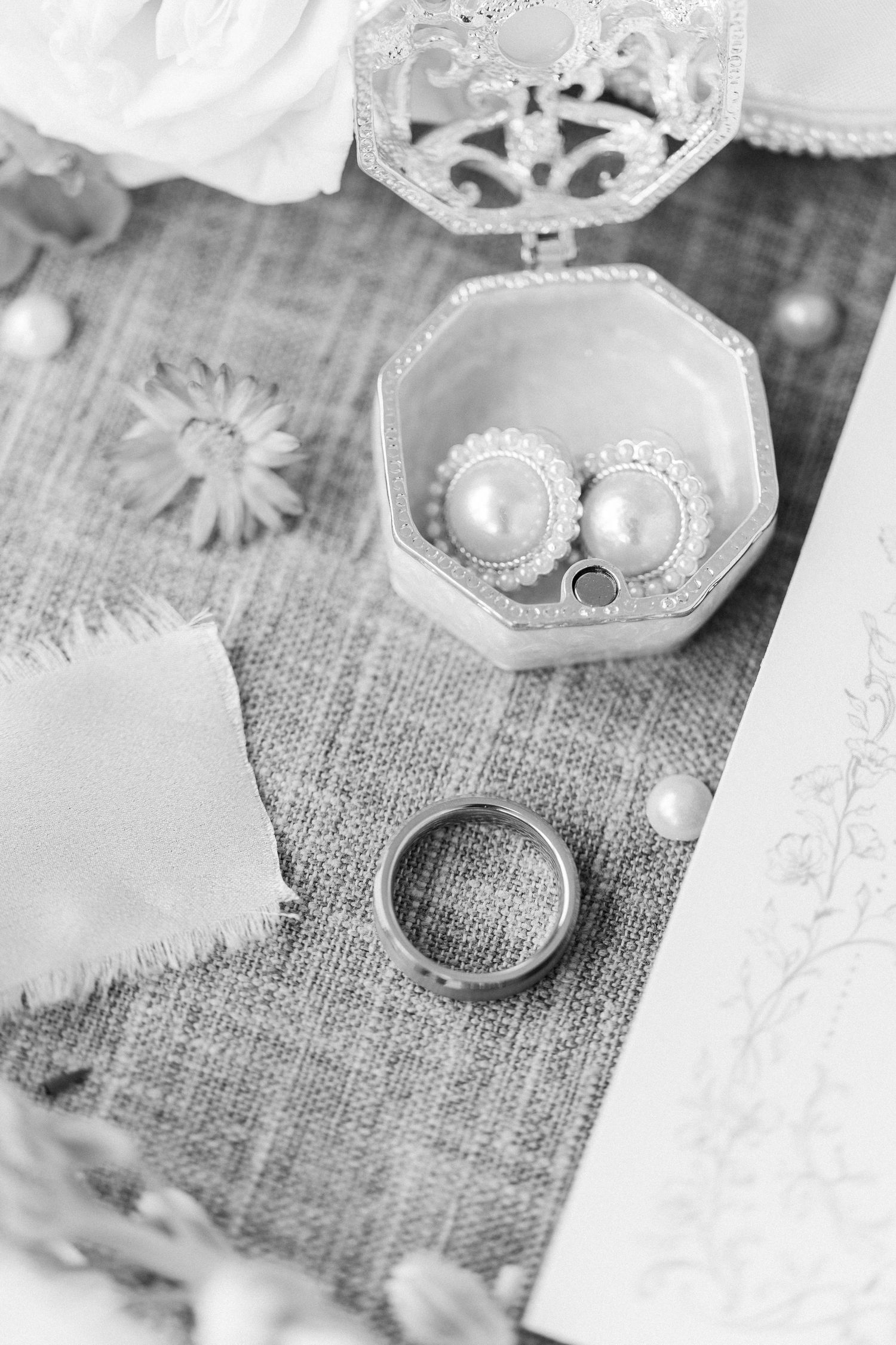 Black and white flat lay of pearl earrings in ornate hexagonal box with wedding band on textured linen.