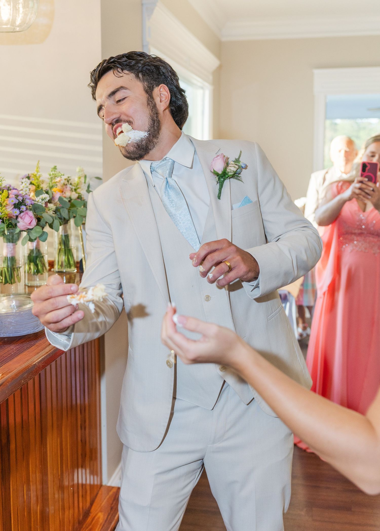 Groom in beige suit playfully eats wedding cake while guests capture the moment at reception.