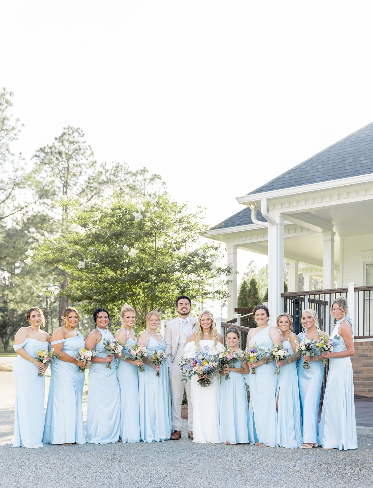 Bride and groom pose with ten bridesmaids in light blue gowns holding colorful bouquets outdoors.