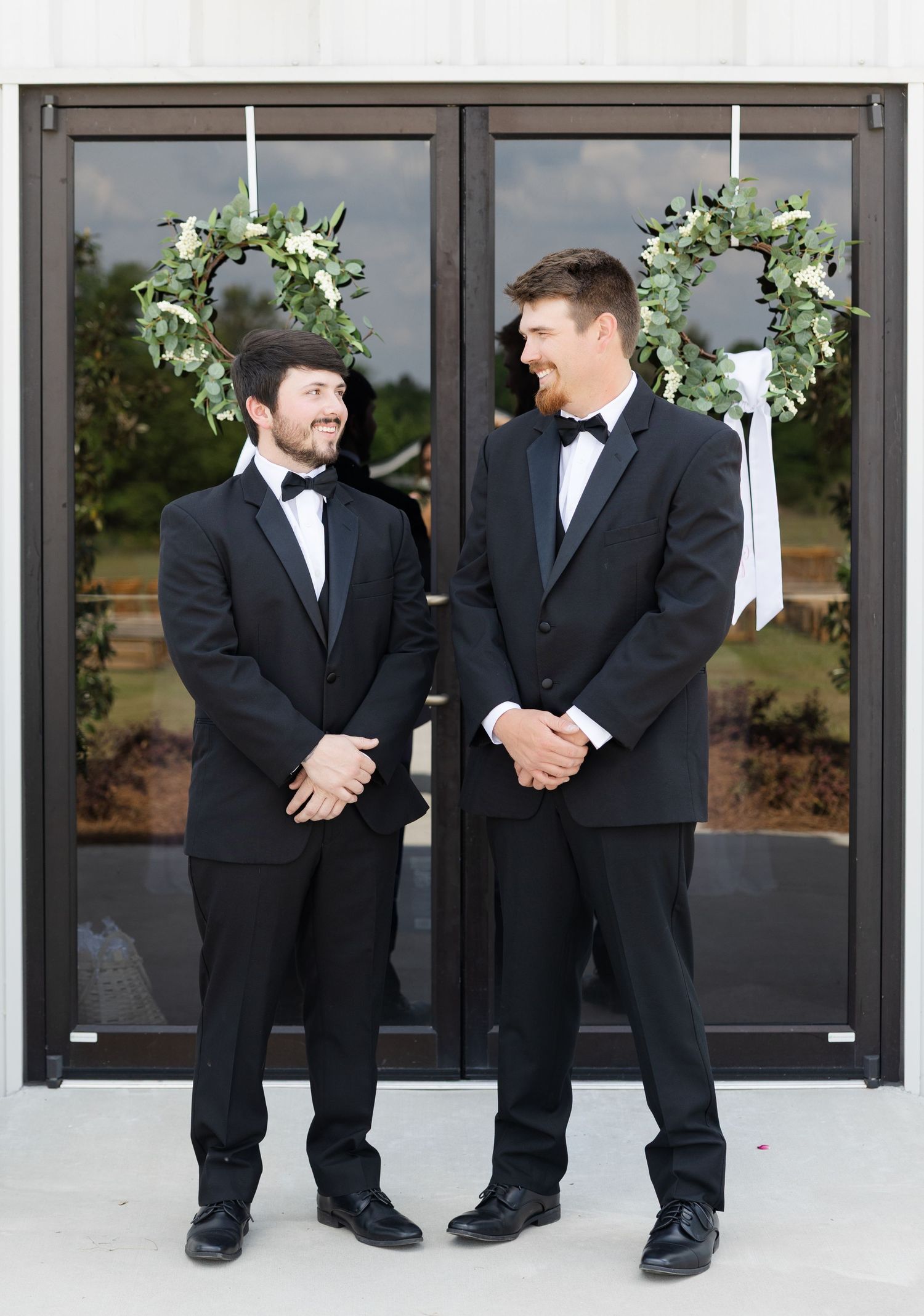 Two groomsmen in black tuxedos standing before doors decorated with greenery wreaths.