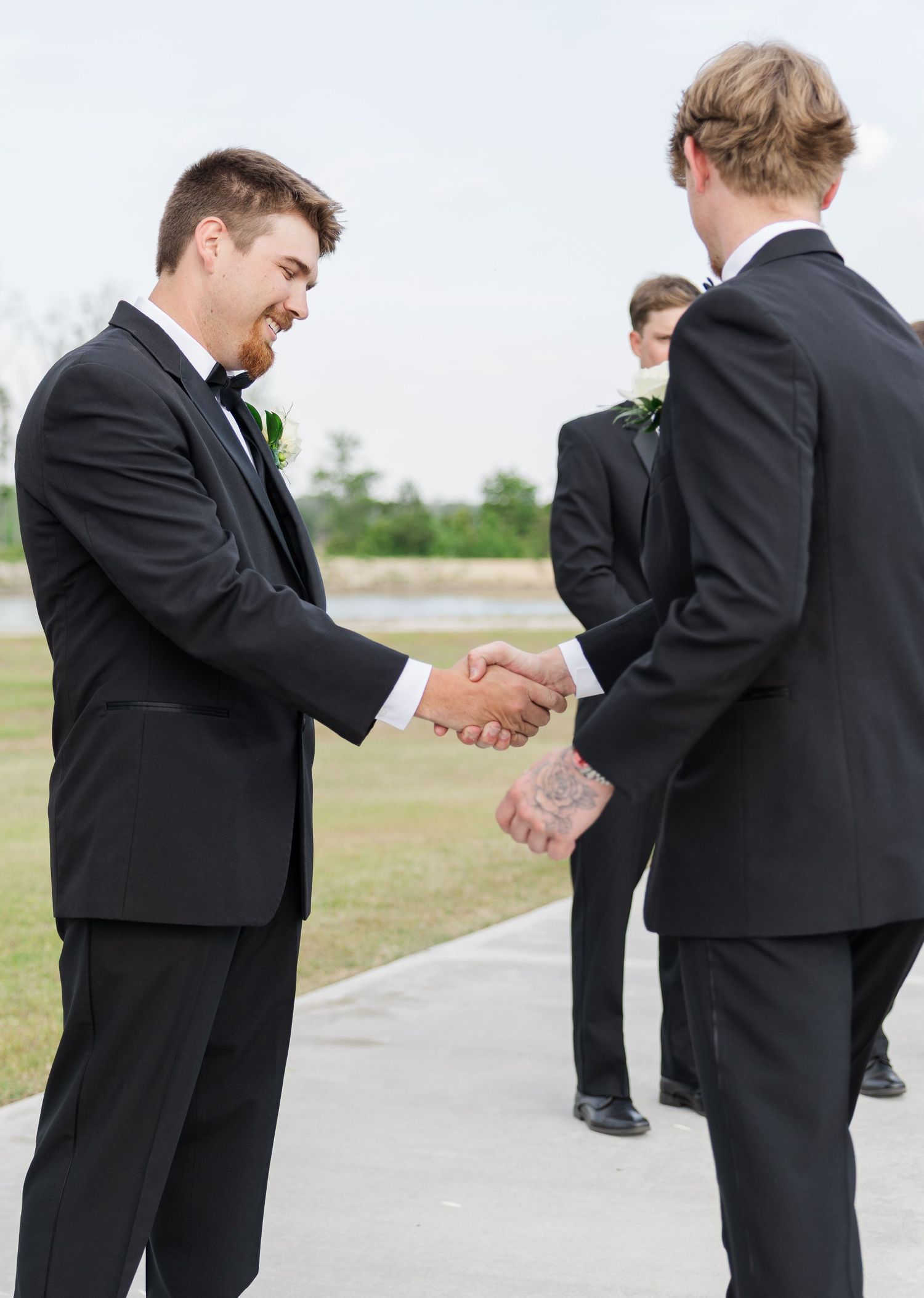 Groom in black tuxedo shakes hands with groomsman outdoors before wedding ceremony.