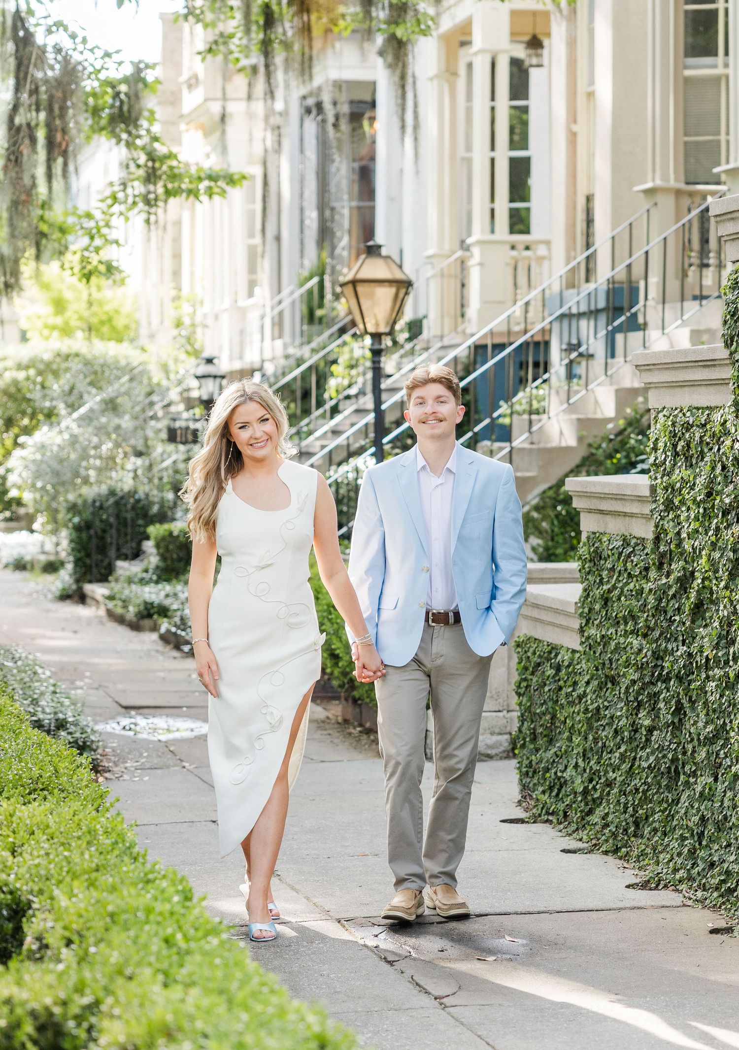 Couple holding hands walking on a sunny sidewalk, woman in white dress, man in light blue blazer and gray pants.