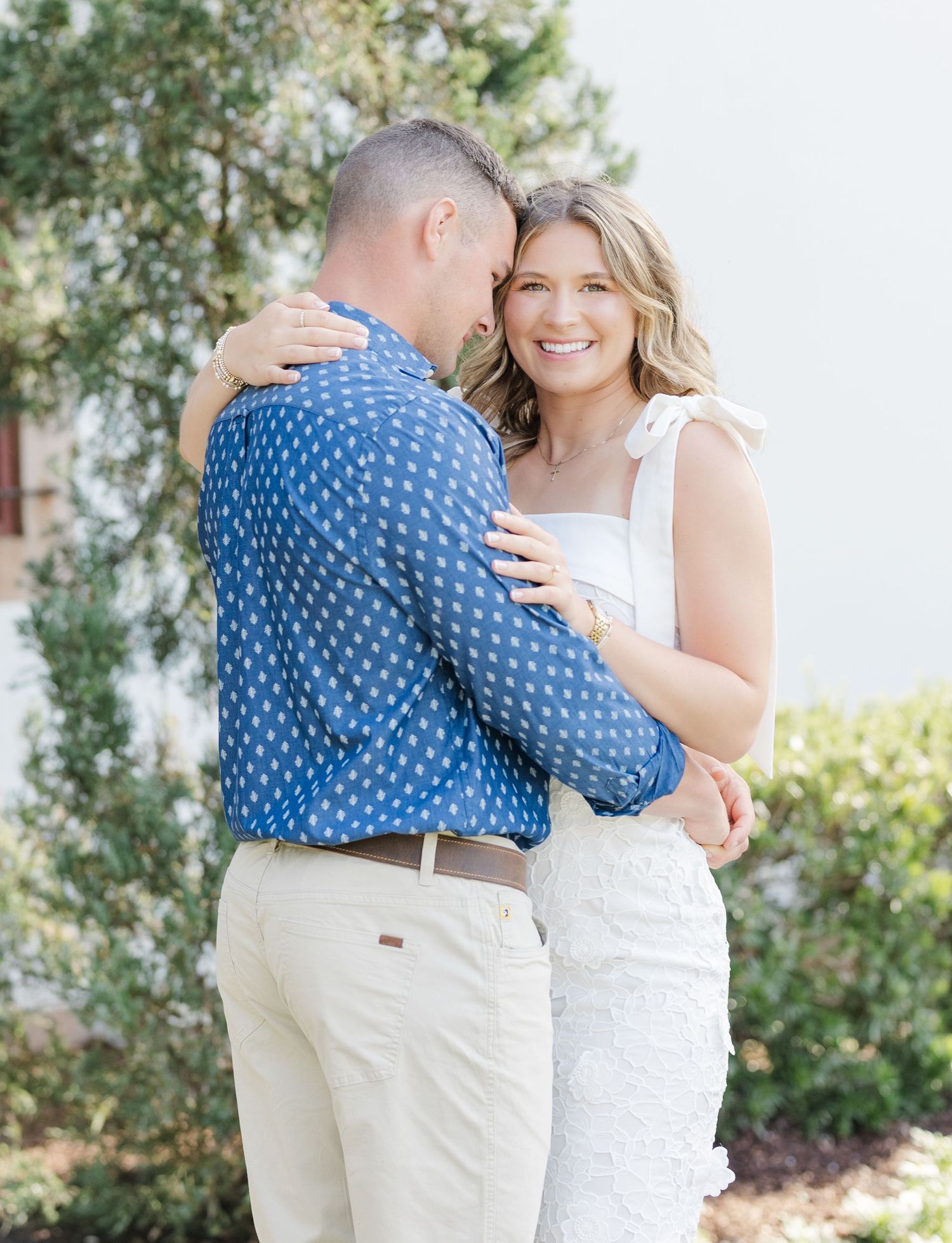 Couple embracing outdoors, woman in white dress smiling, man in blue patterned shirt and khaki pants.