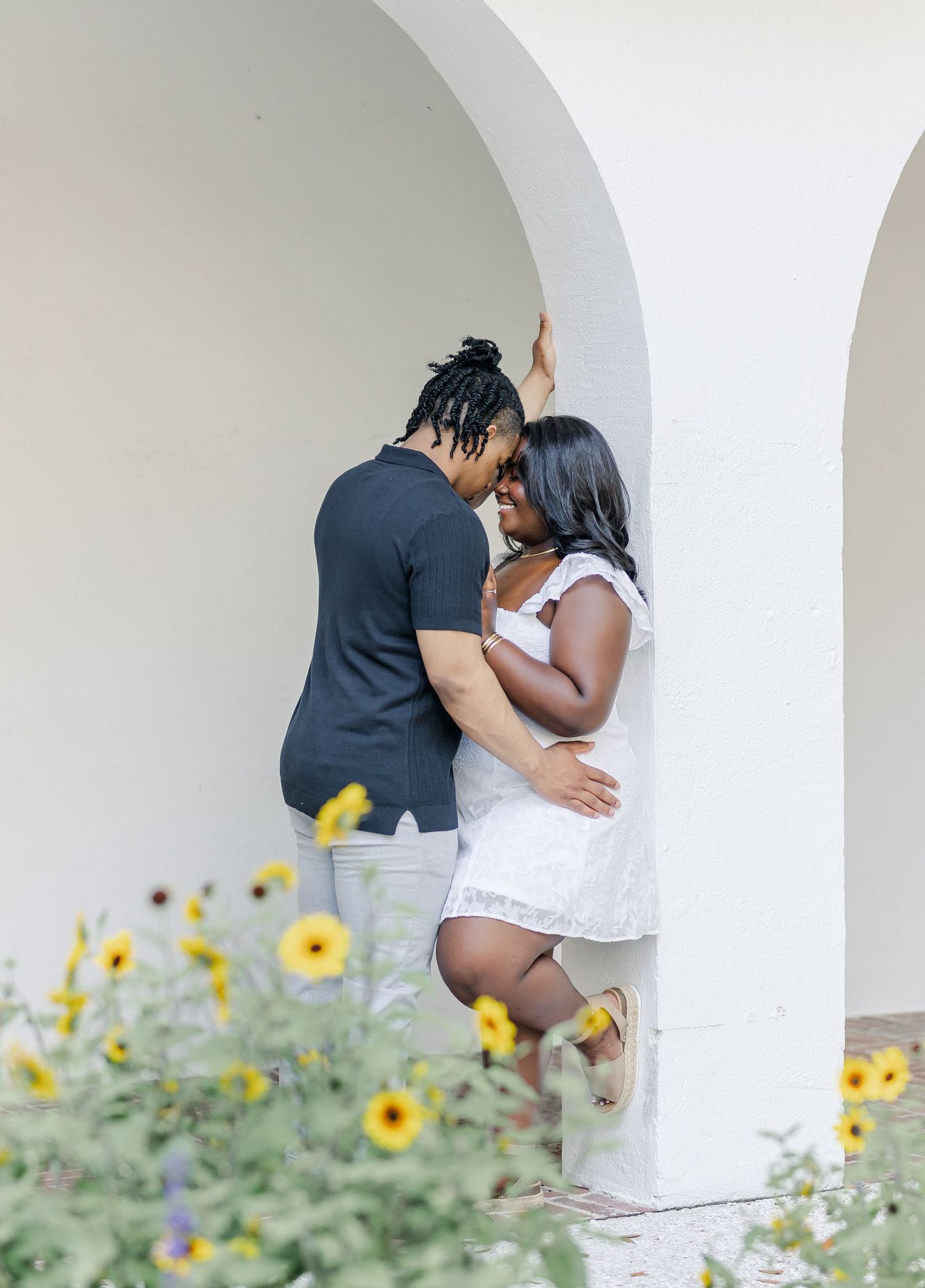 Romantic couple leans close together between white arched columns with yellow sunflowers blooming in foreground.