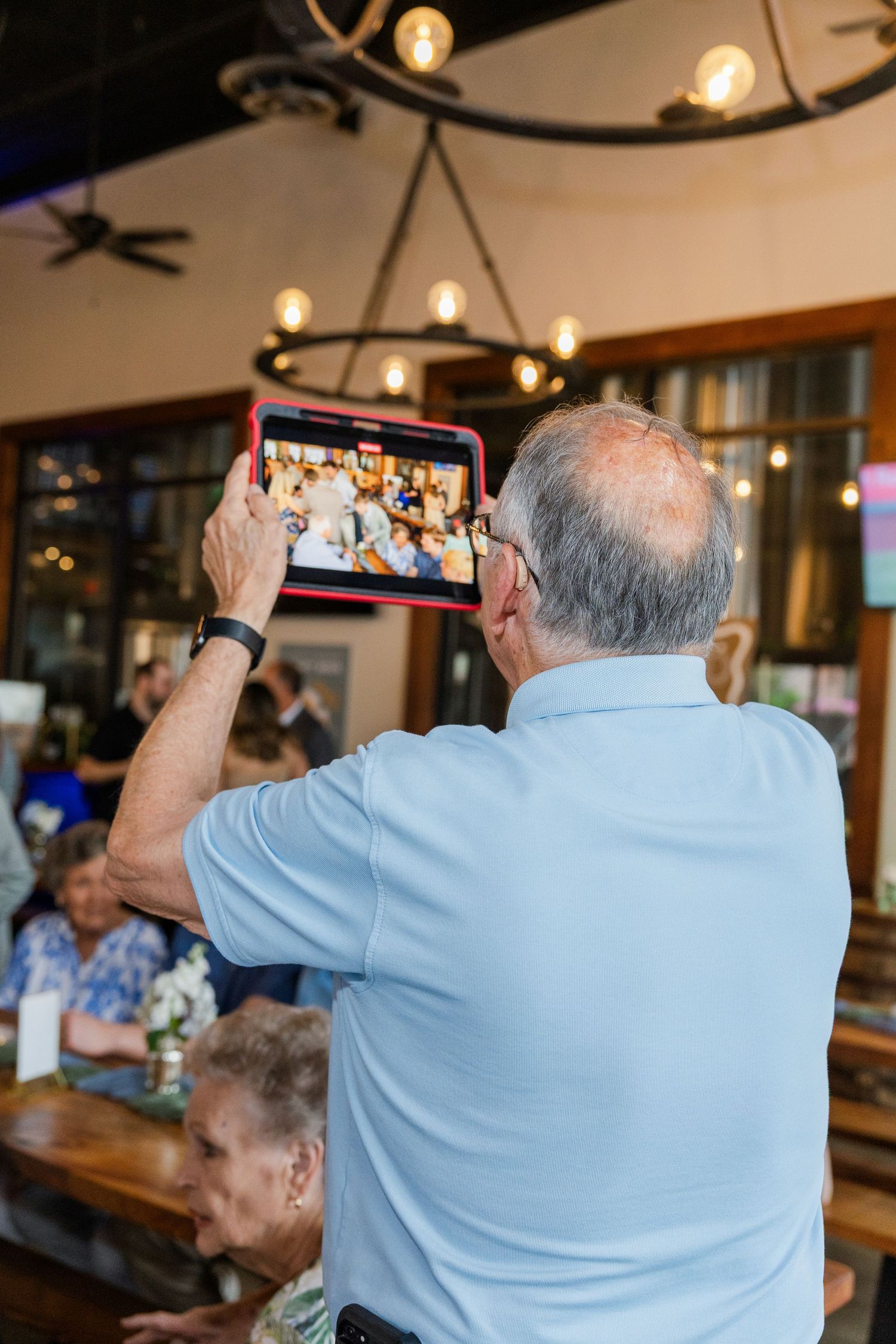 Elderly man photographs a party on a red-framed tablet in a warmly lit restaurant.