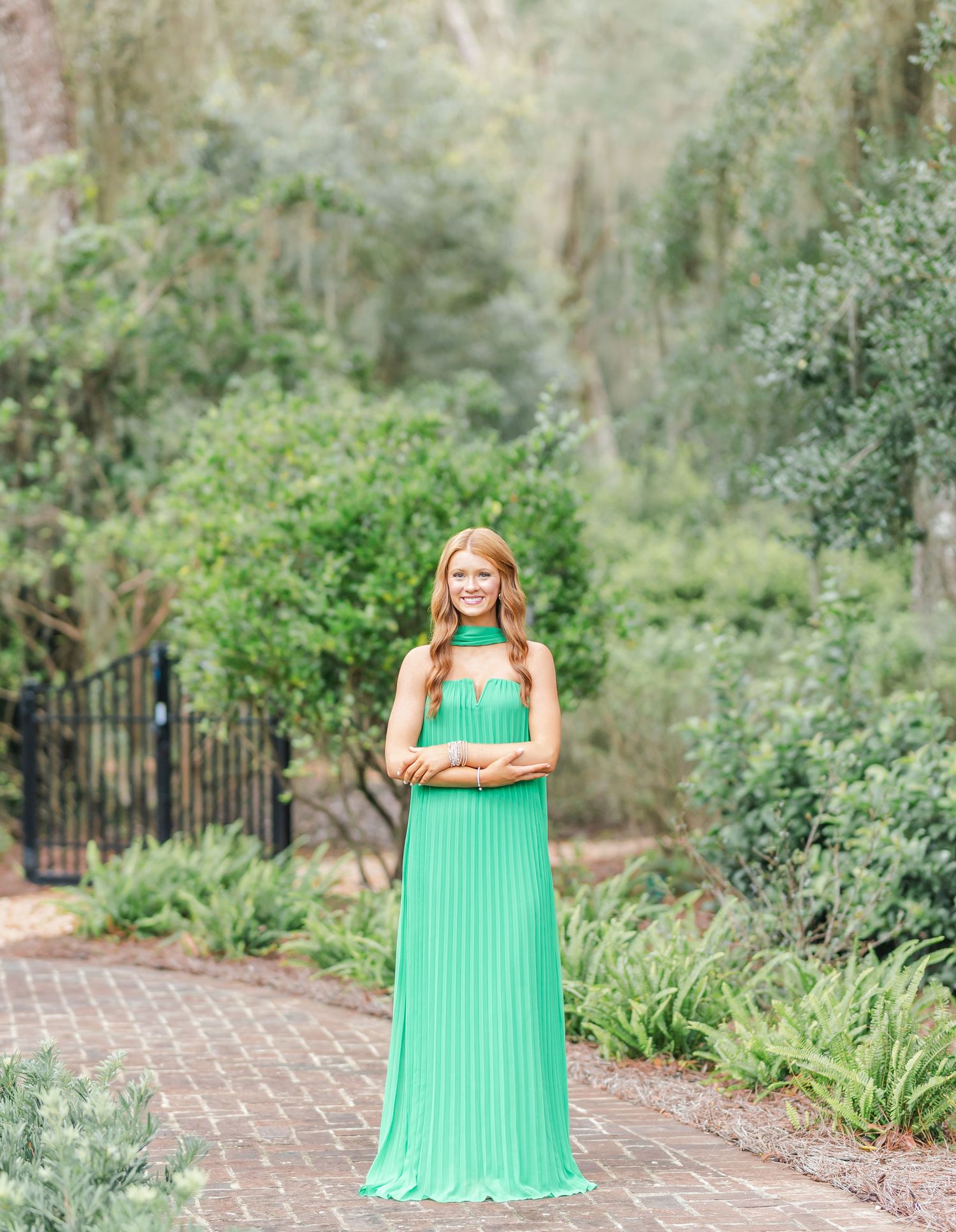A person in a flowing mint green pleated dress stands on a brick pathway surrounded by lush garden foliage.