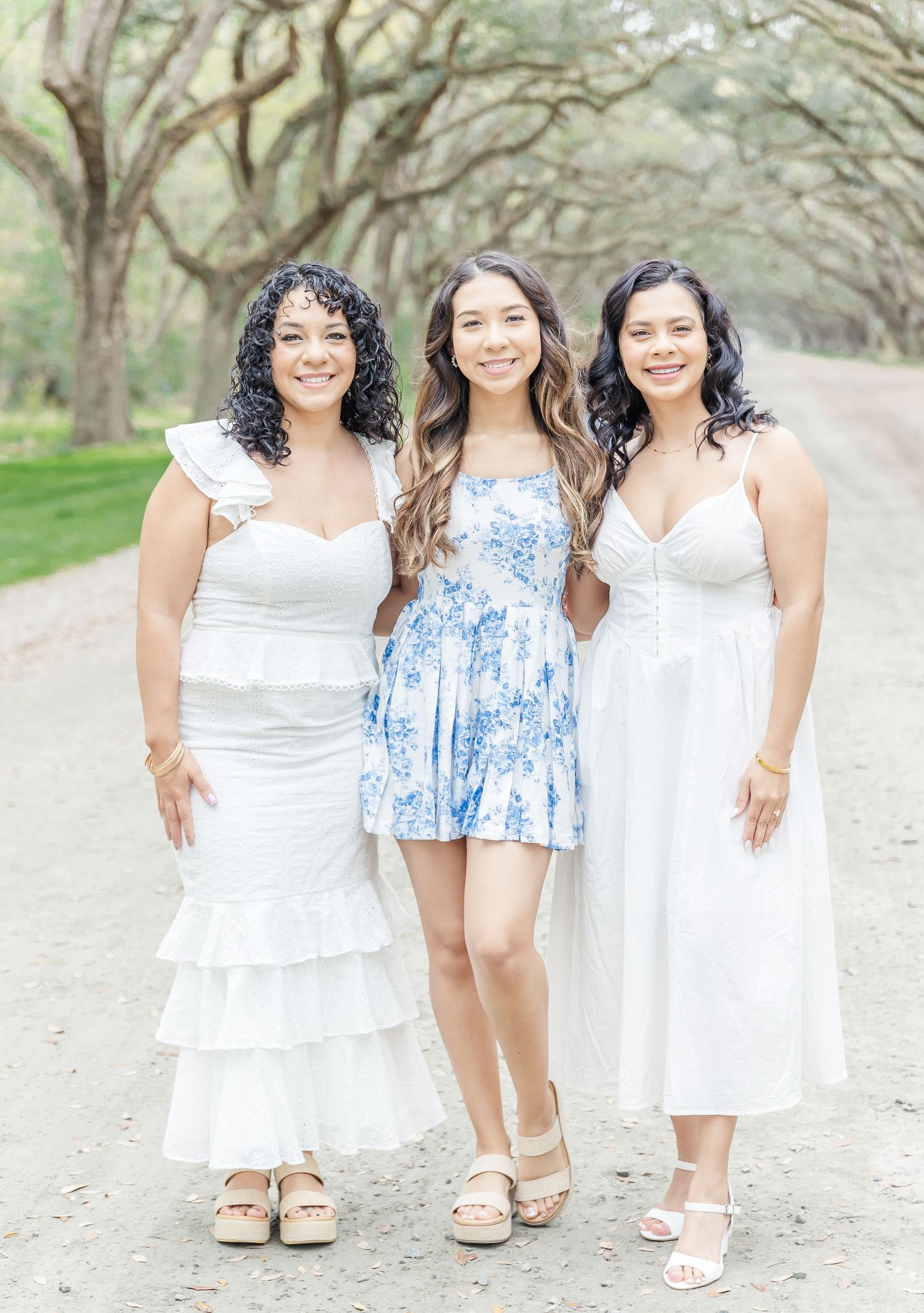 Three women in white and blue dresses standing on a tree-lined path.