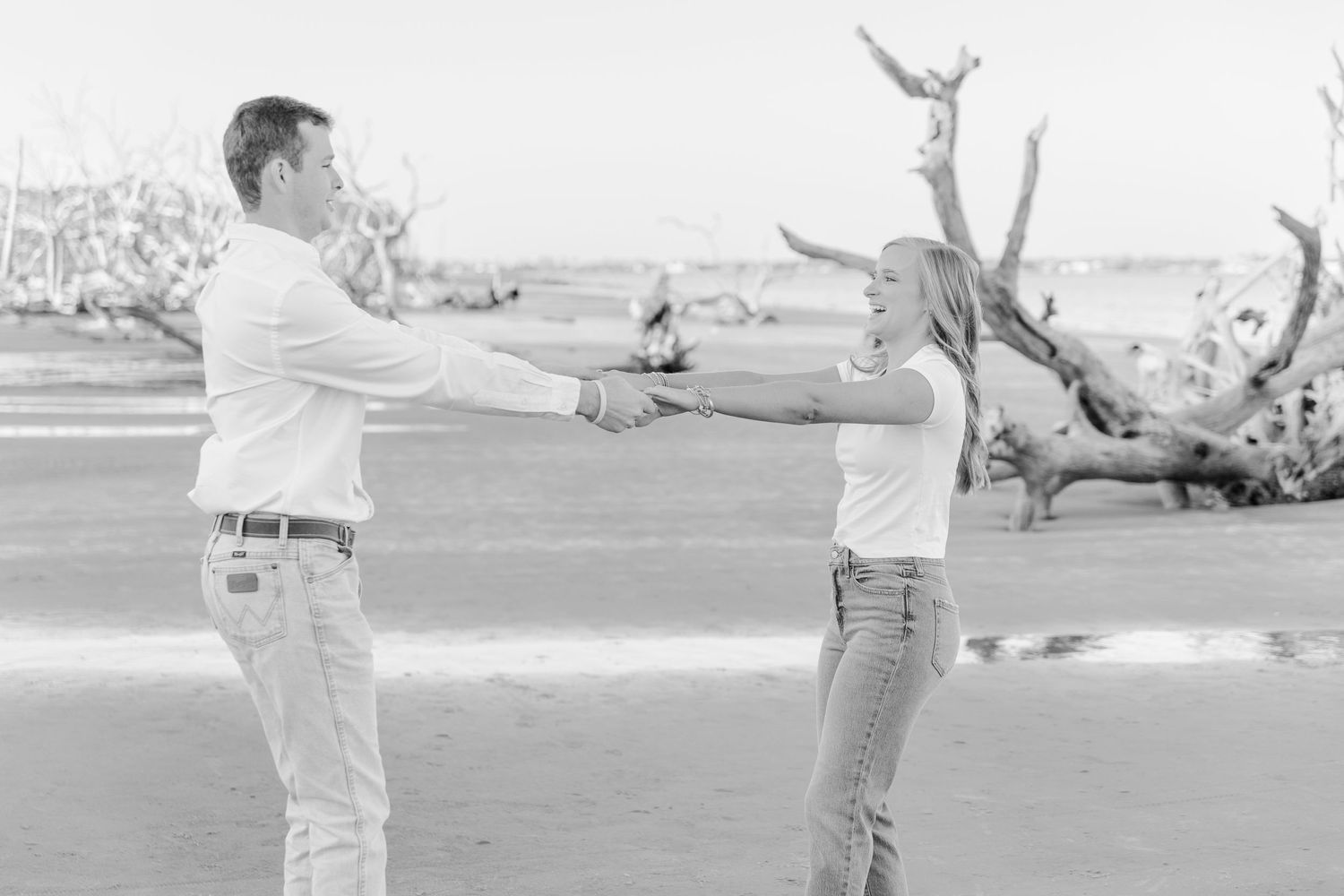 Black and white photo of a couple dancing playfully on a beach with driftwood.