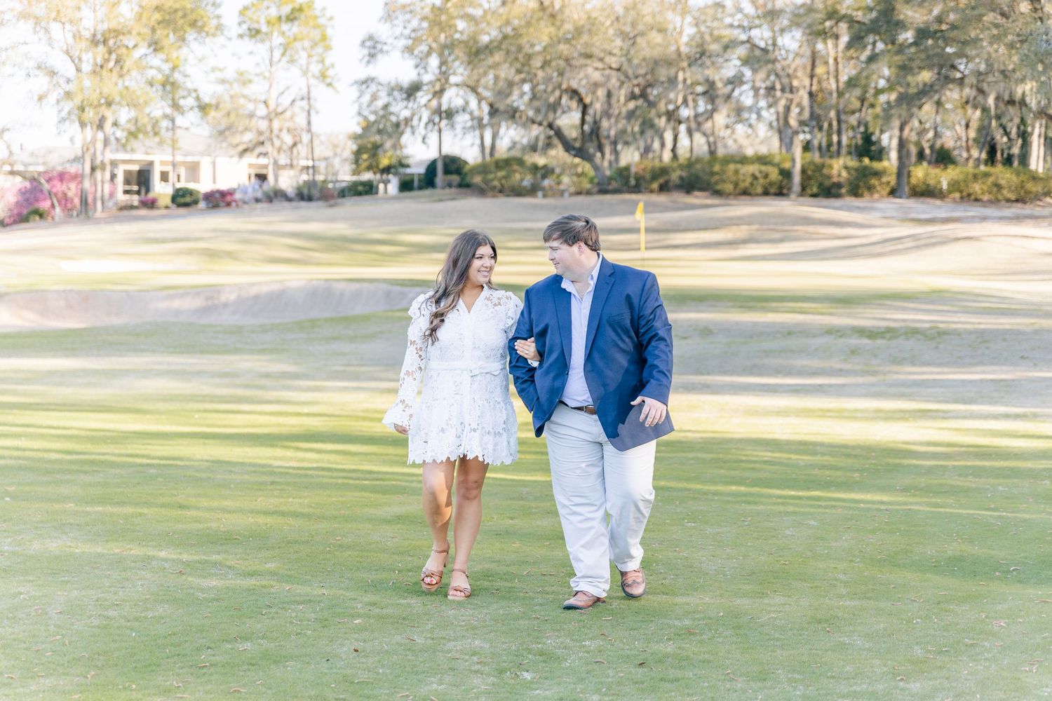 A couple walks hand in hand across a sunny golf course in formal attire.