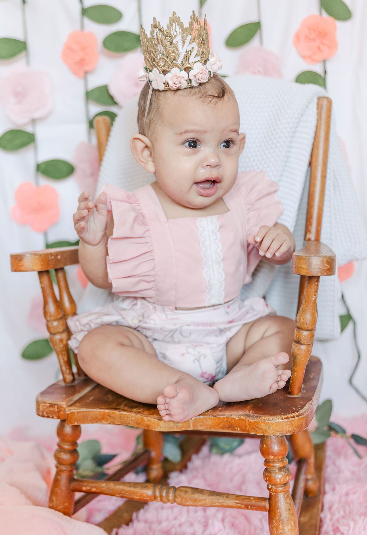 A baby in pink ruffled outfit and gold crown sits in wooden rocking chair against floral backdrop.