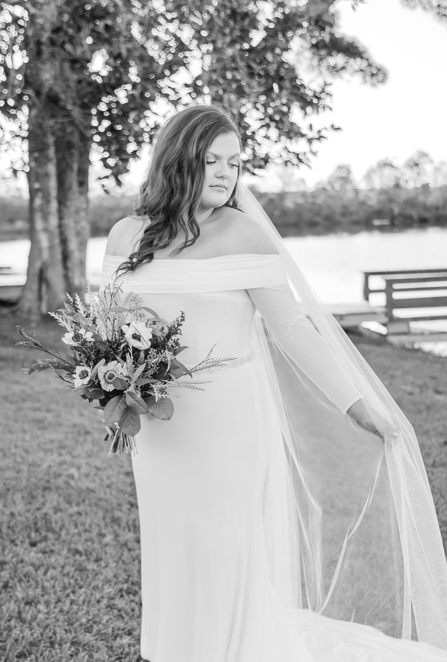 A black and white portrait of a bride in an off-shoulder gown holding a wildflower bouquet outdoors.