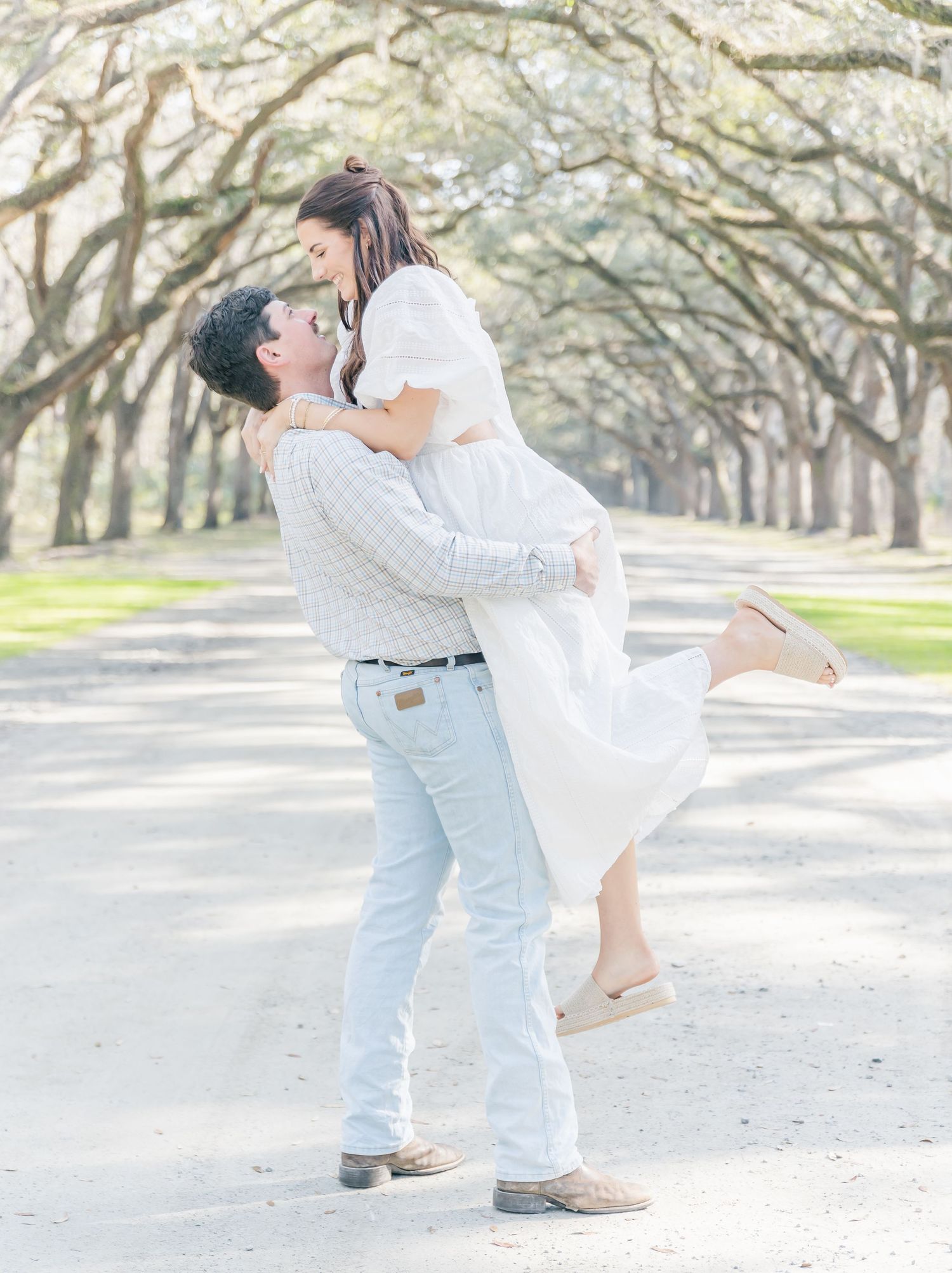 A joyful lift pose between a couple on a tree-lined path in light spring attire.