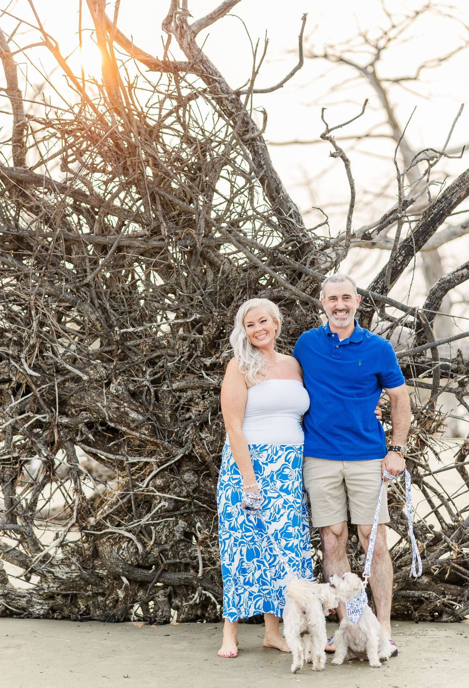 A couple in coordinating blue and white outfits poses with their two small white dogs on a beach.