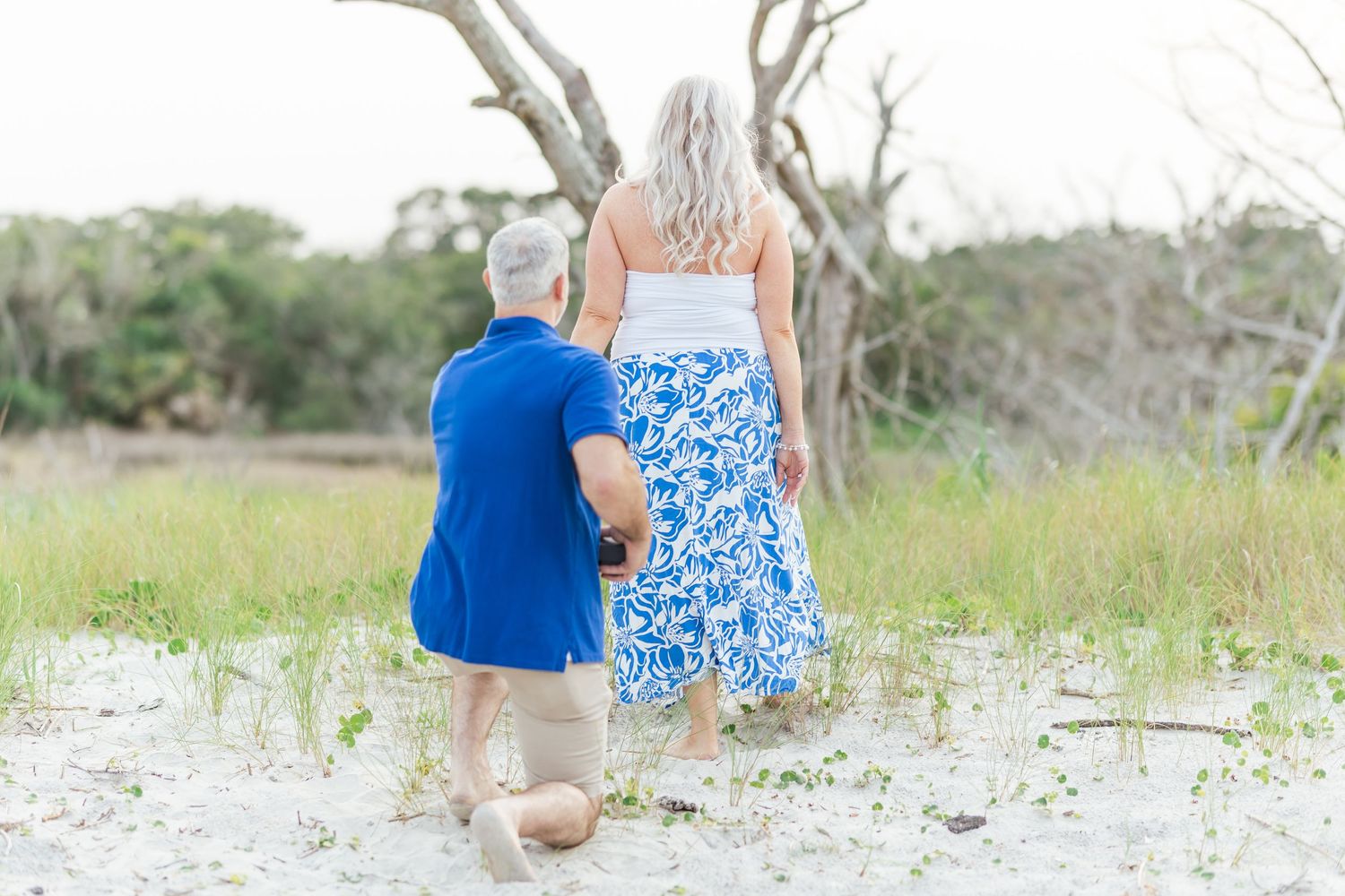 Two people wearing blue clothing sit together on a sandy beach surrounded by coastal vegetation and driftwood.