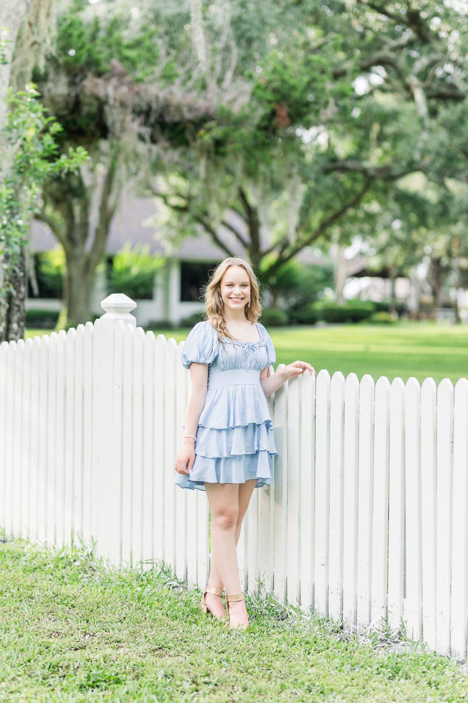 A light blue tiered dress against a white picket fence with Spanish moss draped trees in the background.