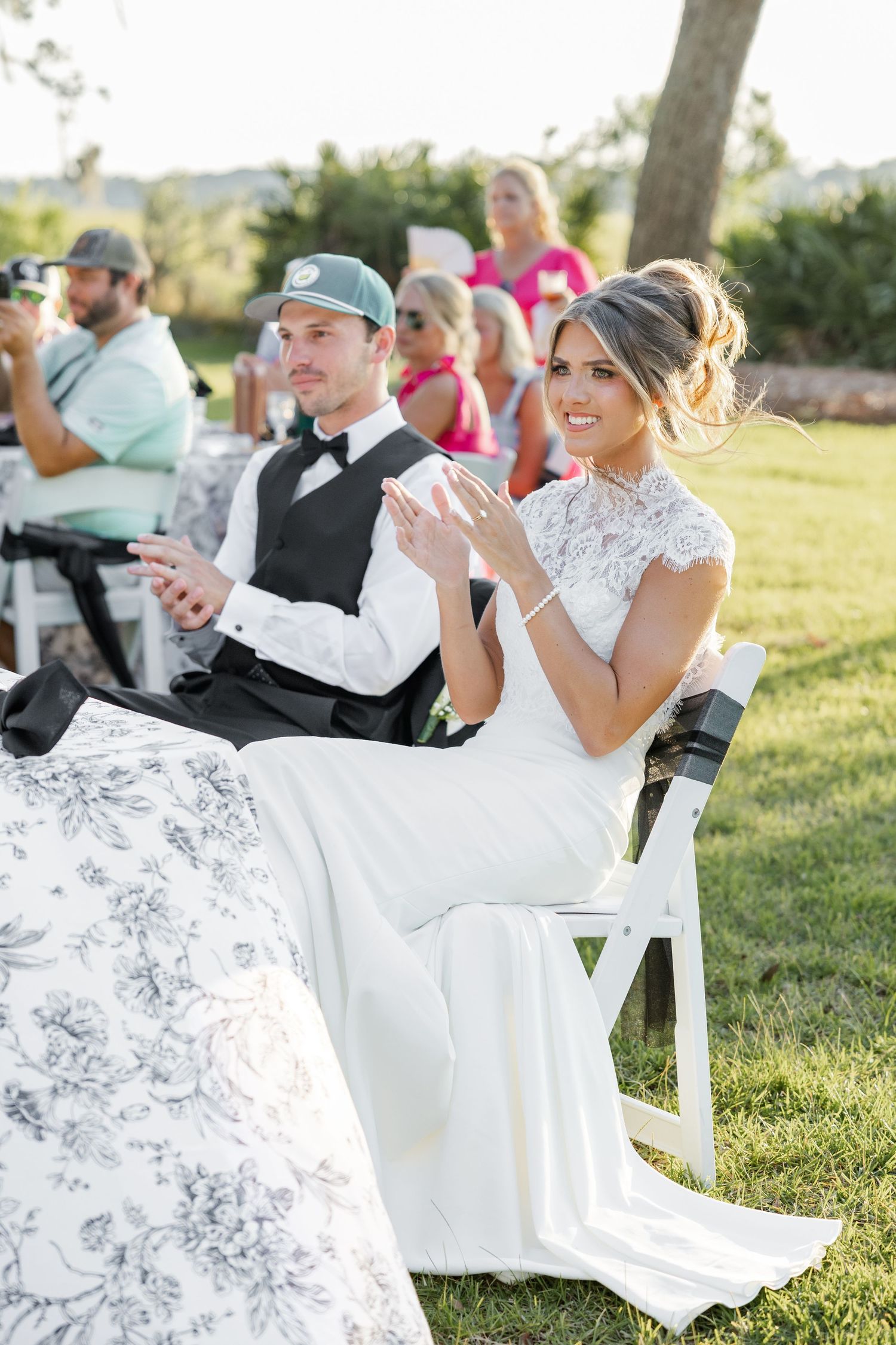 Bride in lace gown and groom clap at outdoor wedding ceremony seated on white chairs.