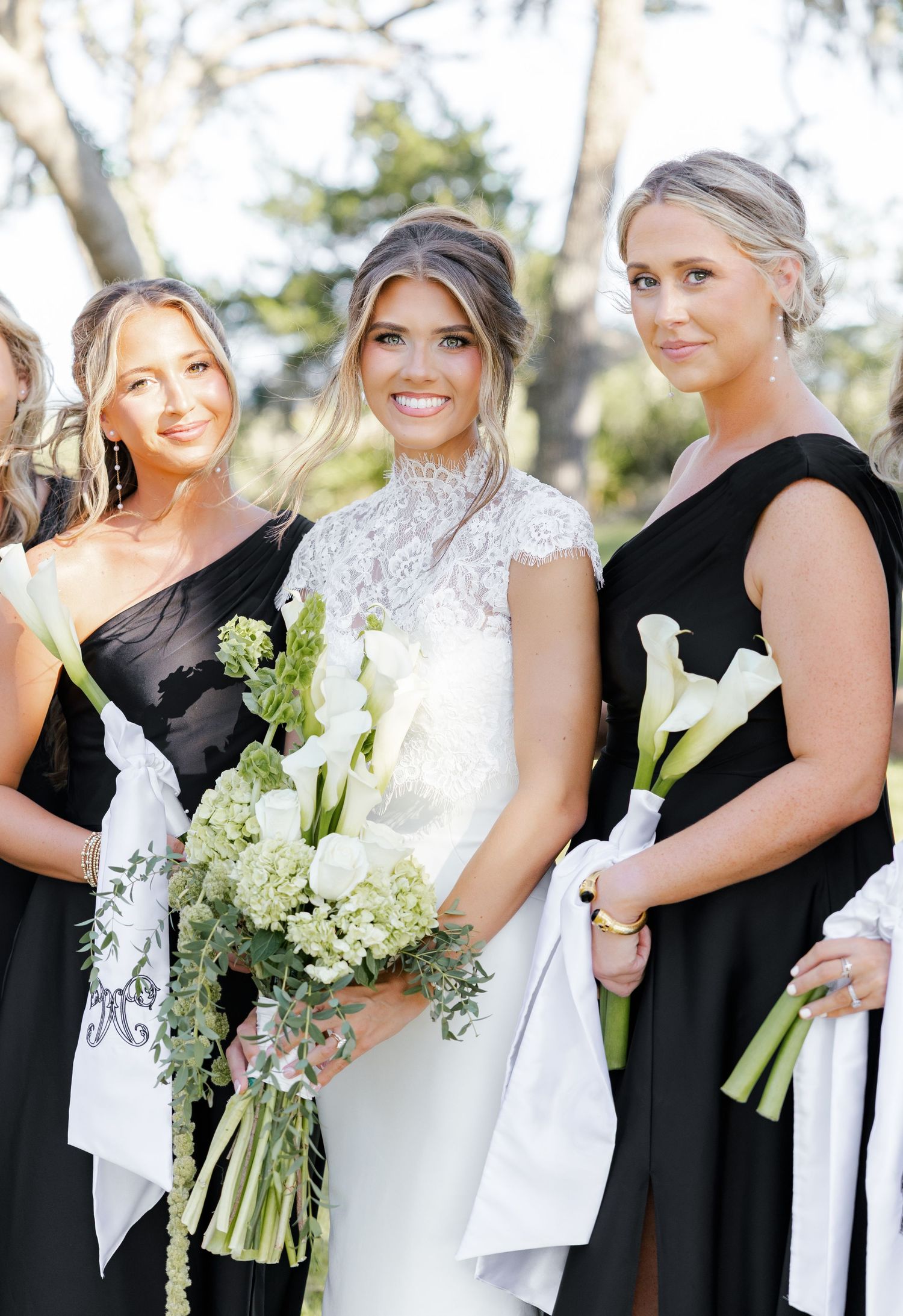 Bride in lace wedding gown smiles with bridesmaids in black dresses holding white calla lily bouquets outdoors.