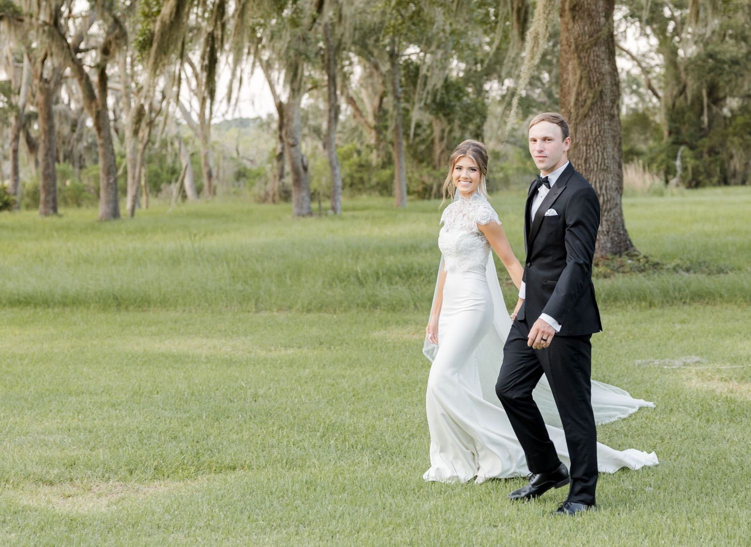 Bride in white gown and groom in black tuxedo posing on lush green lawn with trees in background.