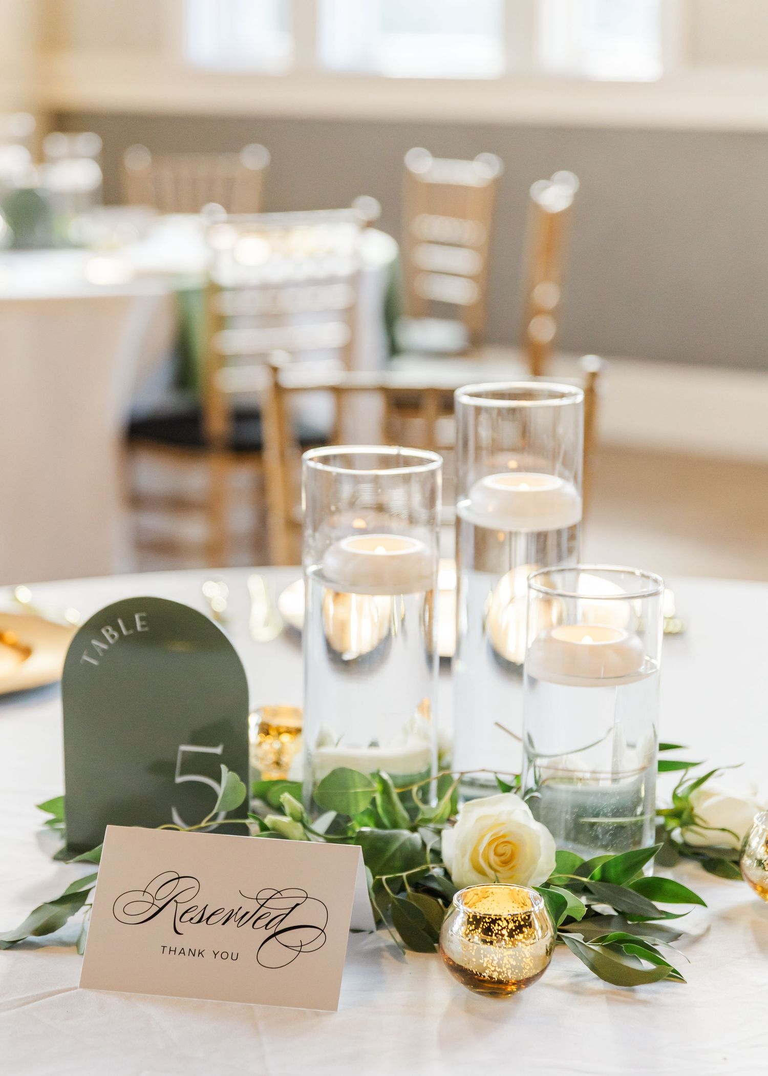 Elegant wedding table centerpiece with floating candles, white roses, greenery, and reserved sign at reception.