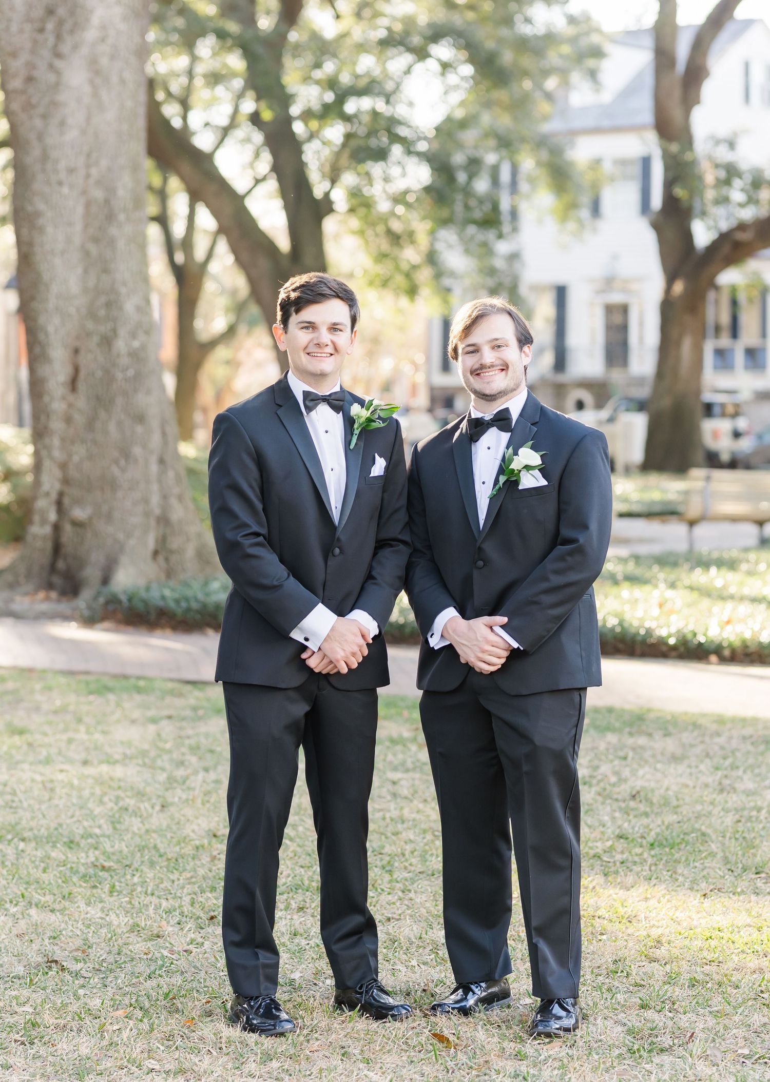 Two groomsmen in classic black tuxedos with white boutonnieres smile together outdoors at a wedding celebration.