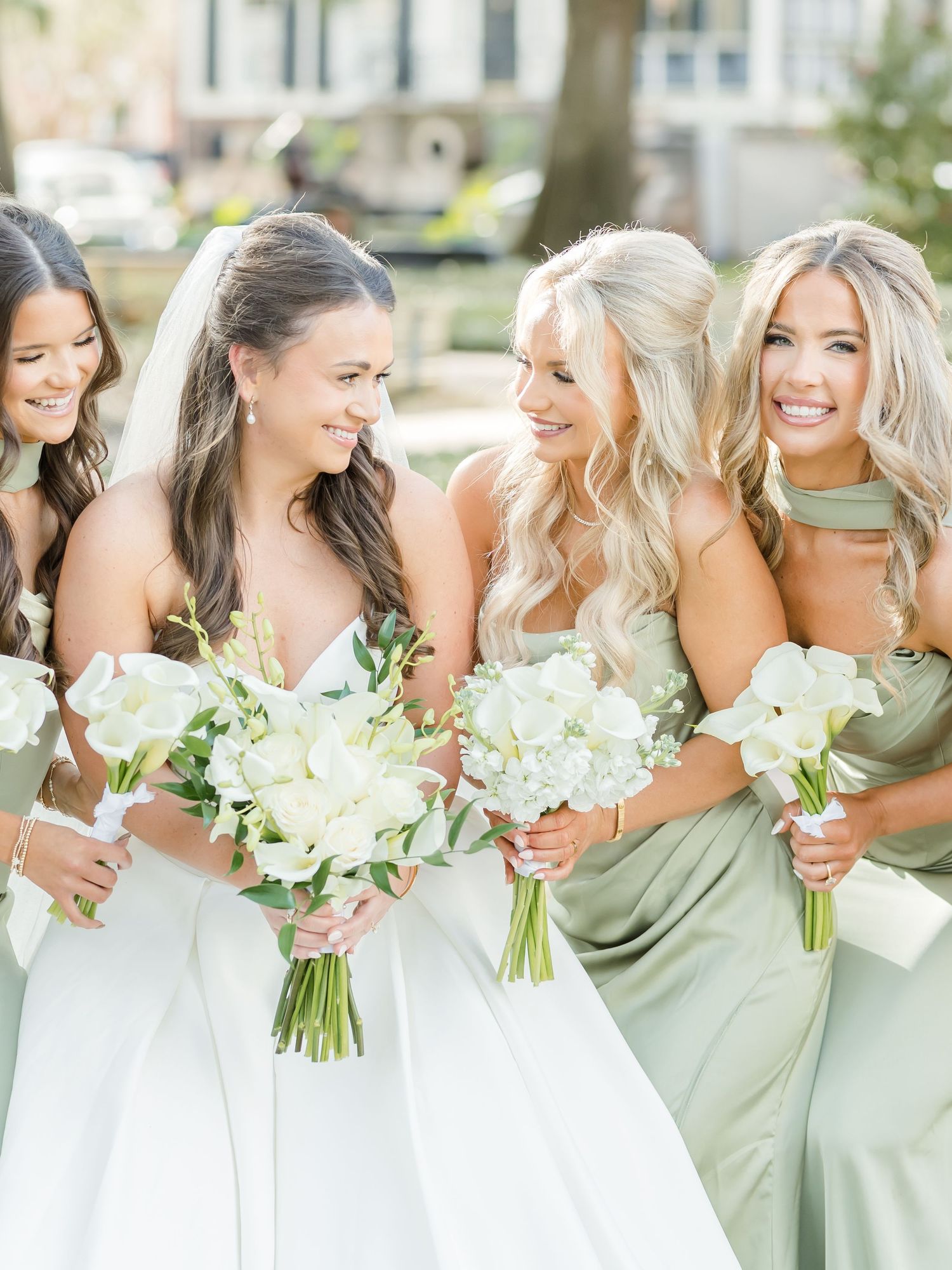 Bride in white strapless gown laughs with three bridesmaids in sage green dresses, all holding white floral bouquets.