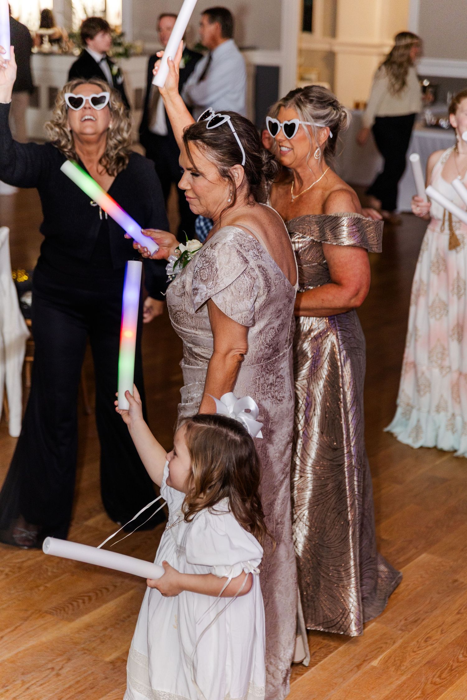 Wedding guests dance with glow sticks, wearing heart-shaped sunglasses, including a flower girl in white dress.
