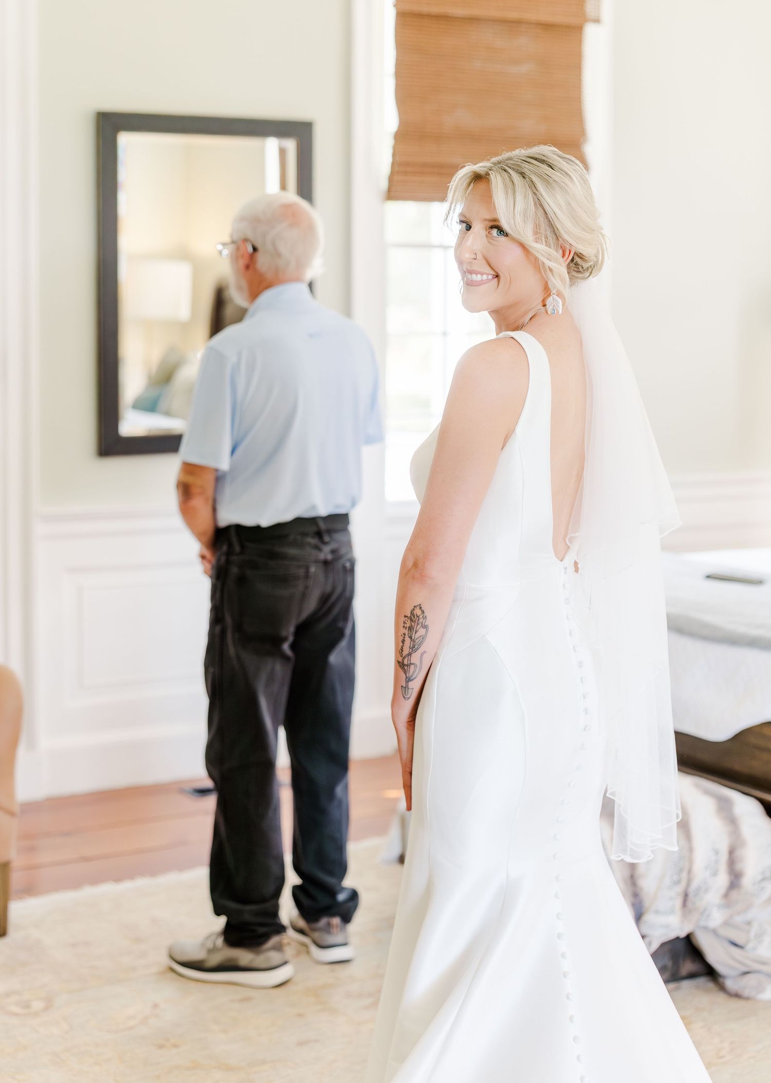 Bride in backless white gown smiles during first look moment with father indoors.