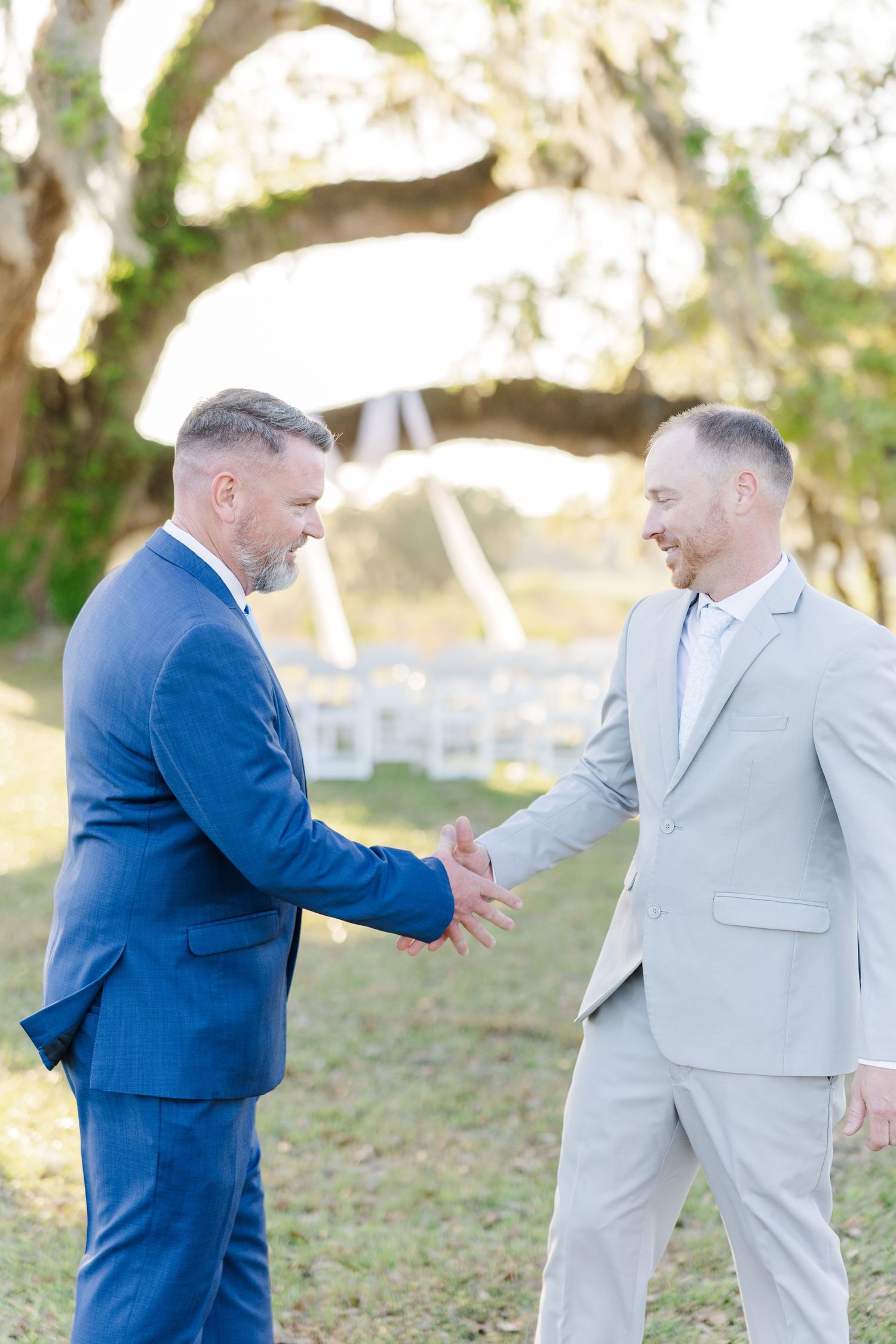 Two men in suits shake hands warmly outdoors near a wedding ceremony setup under a large oak tree.