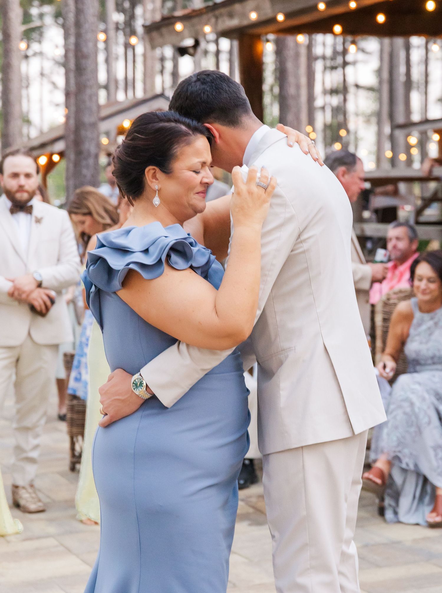 Groom in beige suit shares emotional mother-son dance at outdoor wedding reception surrounded by guests.