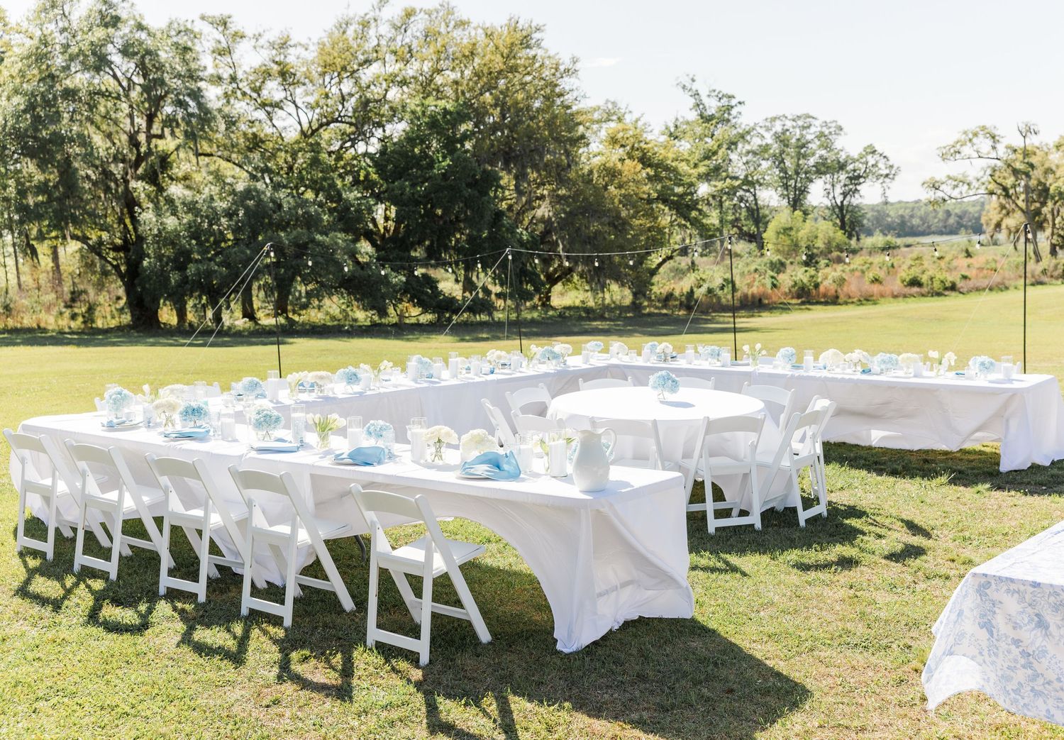 Elegant outdoor wedding reception tables set with white linens and blue accents on a sunny green lawn.