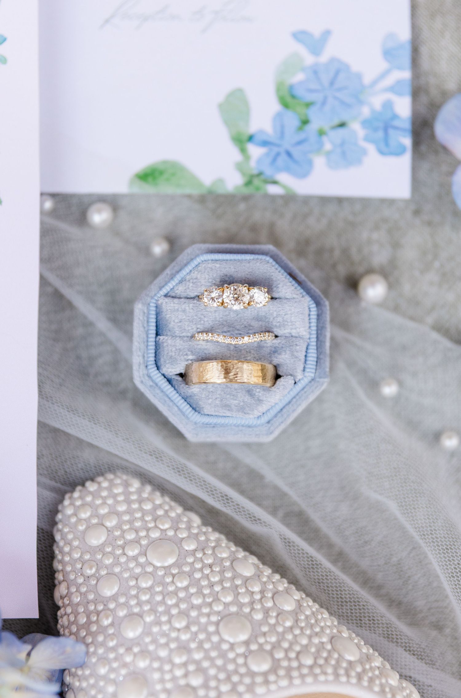 Three gold wedding rings displayed in a blue velvet octagonal ring box with pearl-adorned bridal shoes nearby.