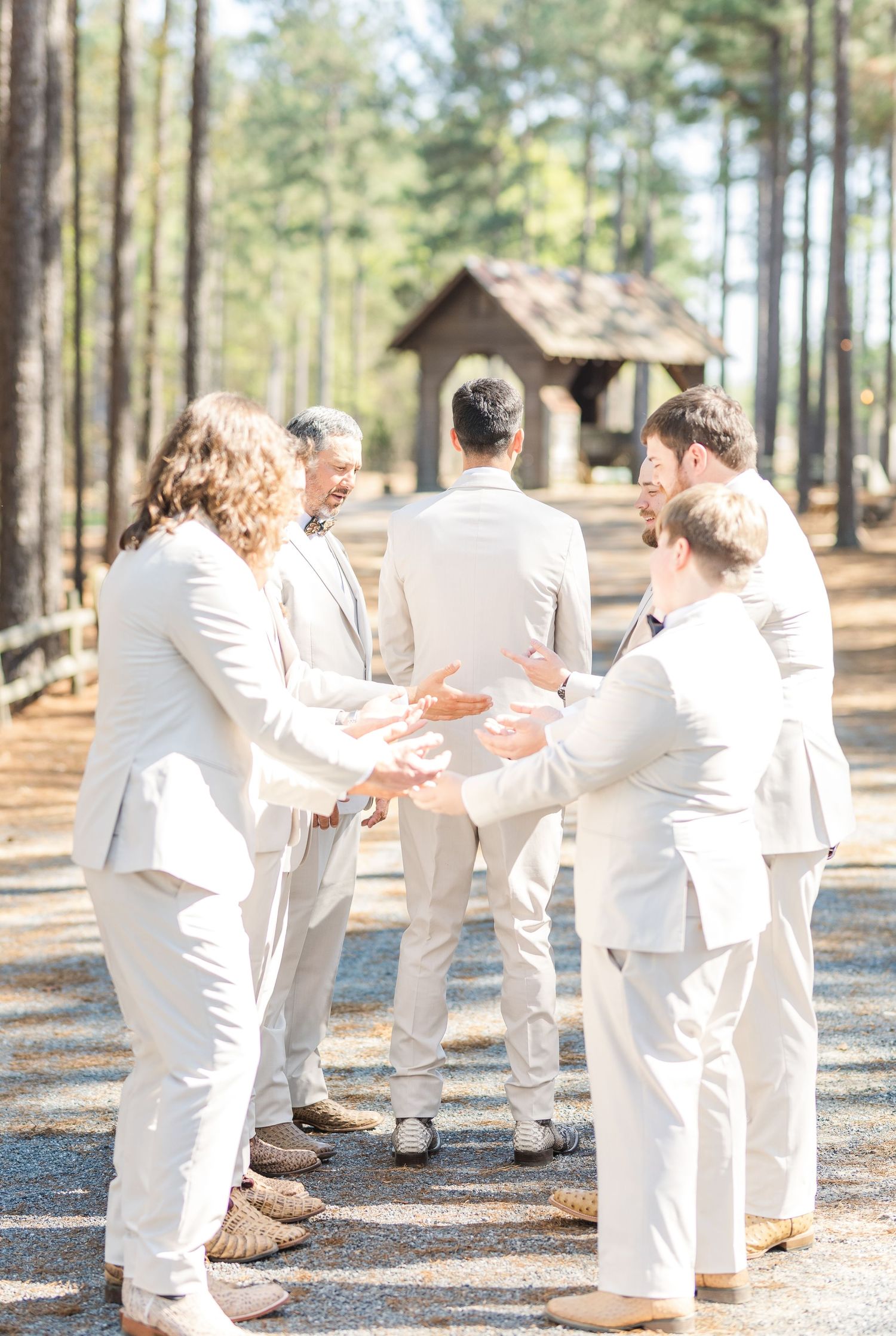 Groomsmen in cream suits sharing a prayer circle at outdoor rustic wedding venue.