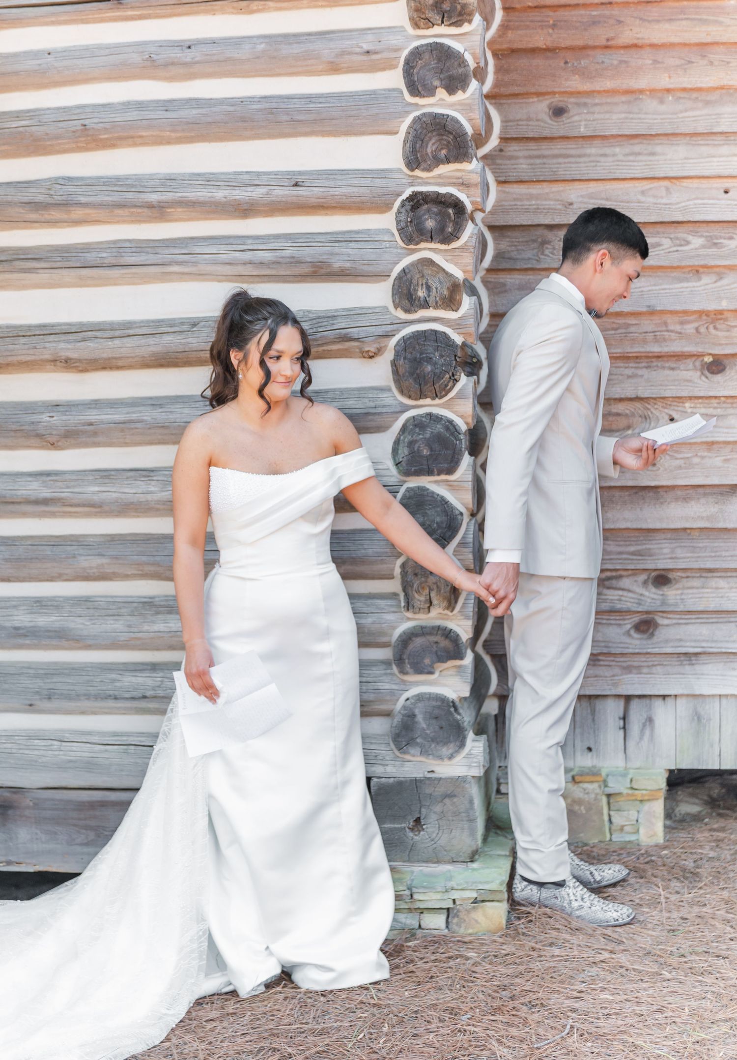 Bride in off-shoulder white gown holds grooms hand during first look against rustic log cabin wall.