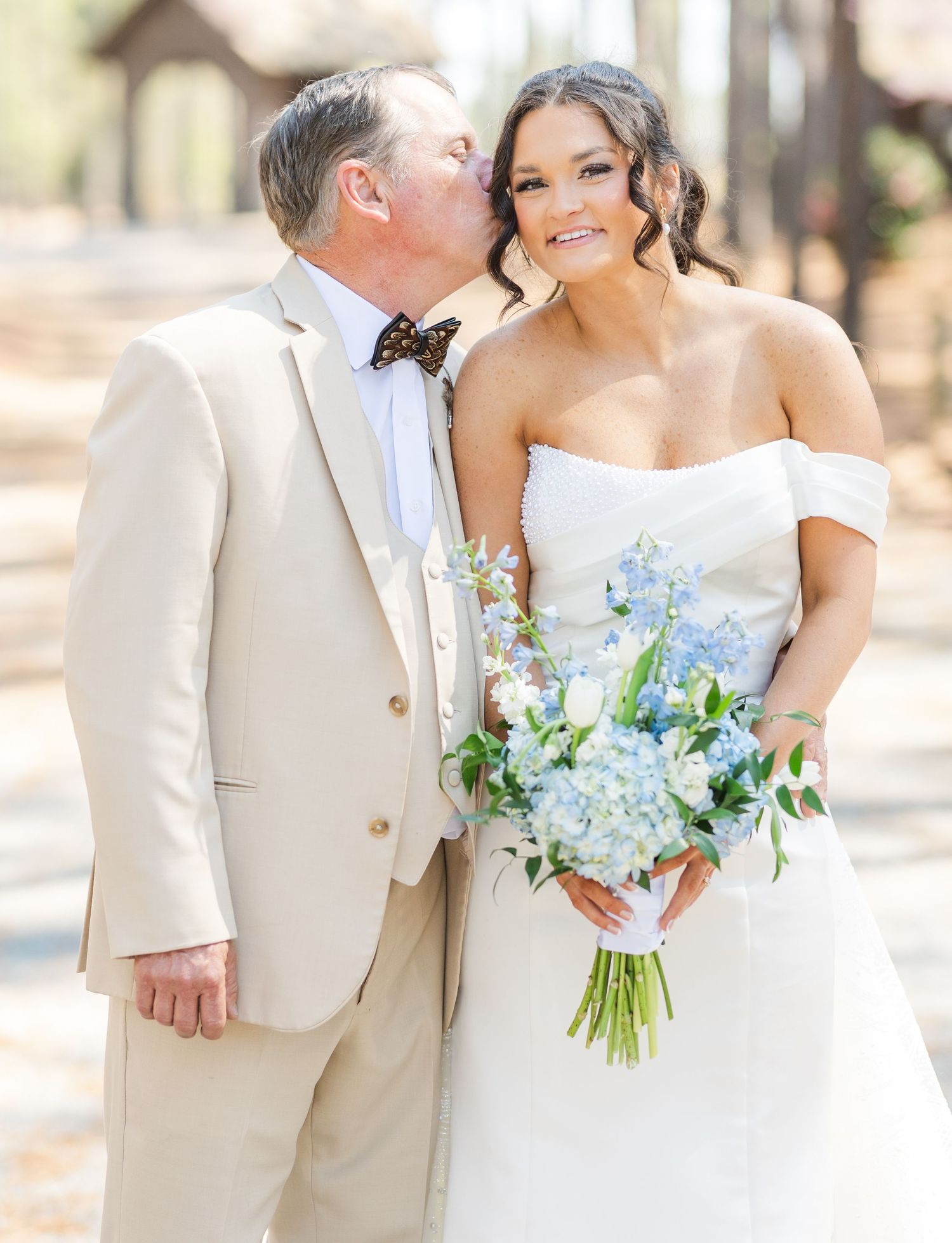 Bride in off-shoulder gown smiles as older man in ivory suit kisses her cheek outdoors.