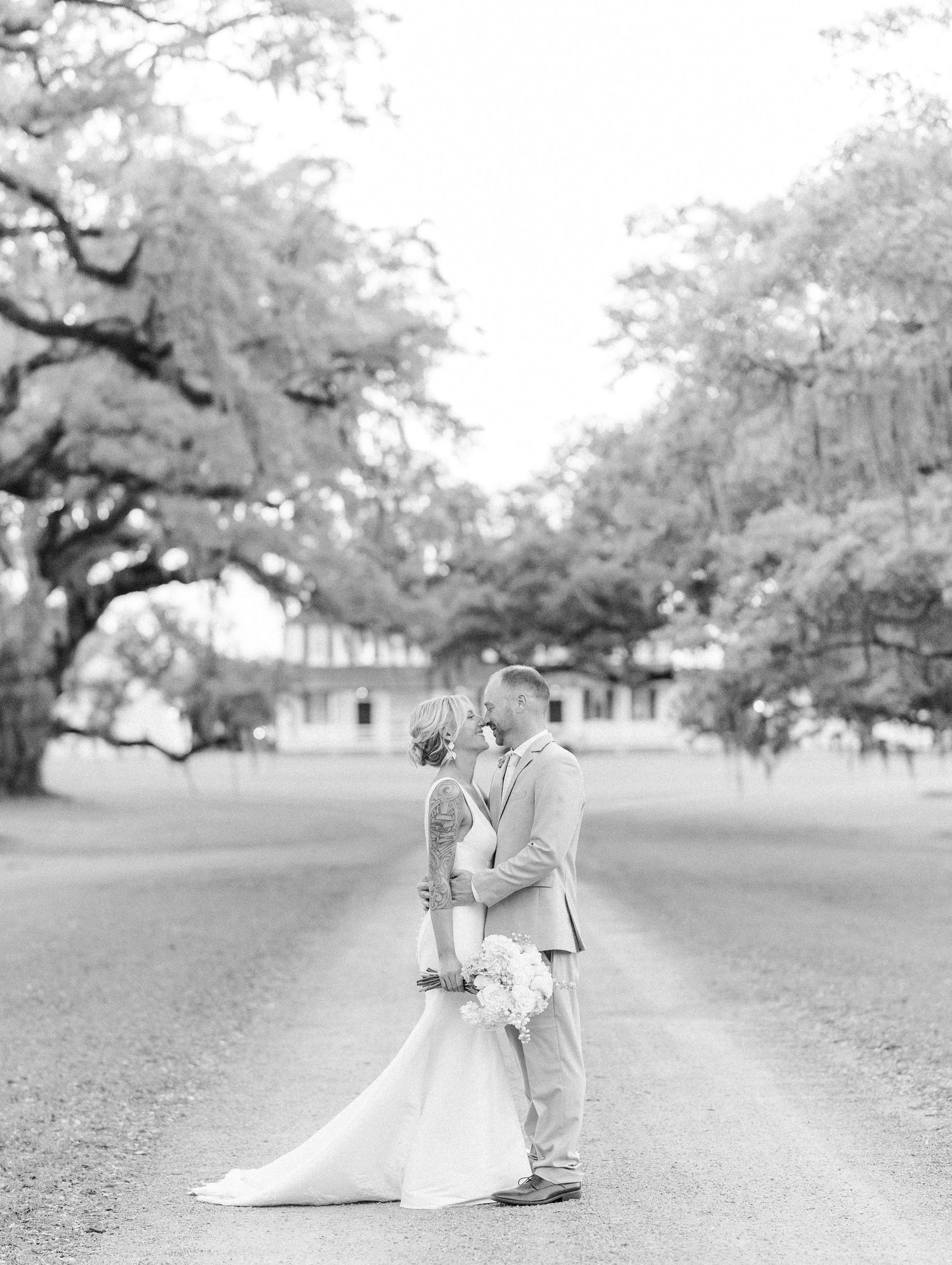 Black and white romantic photo of bride and groom sharing a kiss on a tree-lined path at wedding venue.