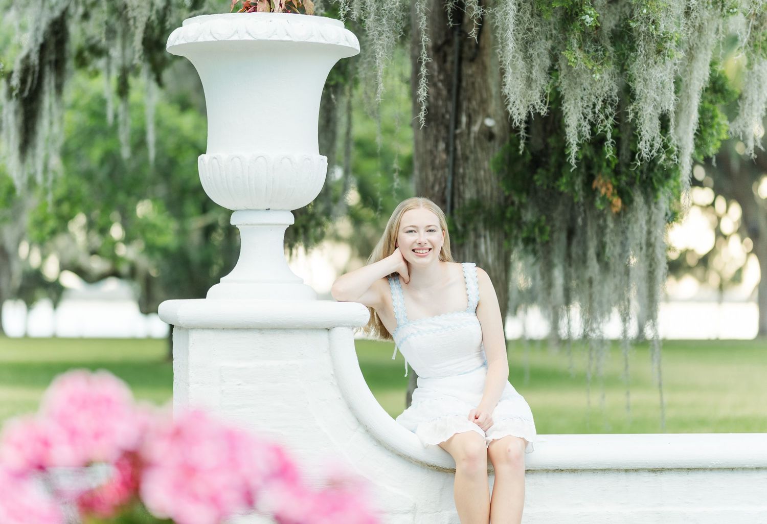 A serene garden scene featuring a white stone urn and Spanish moss draped trees with pink flowers in the foreground.