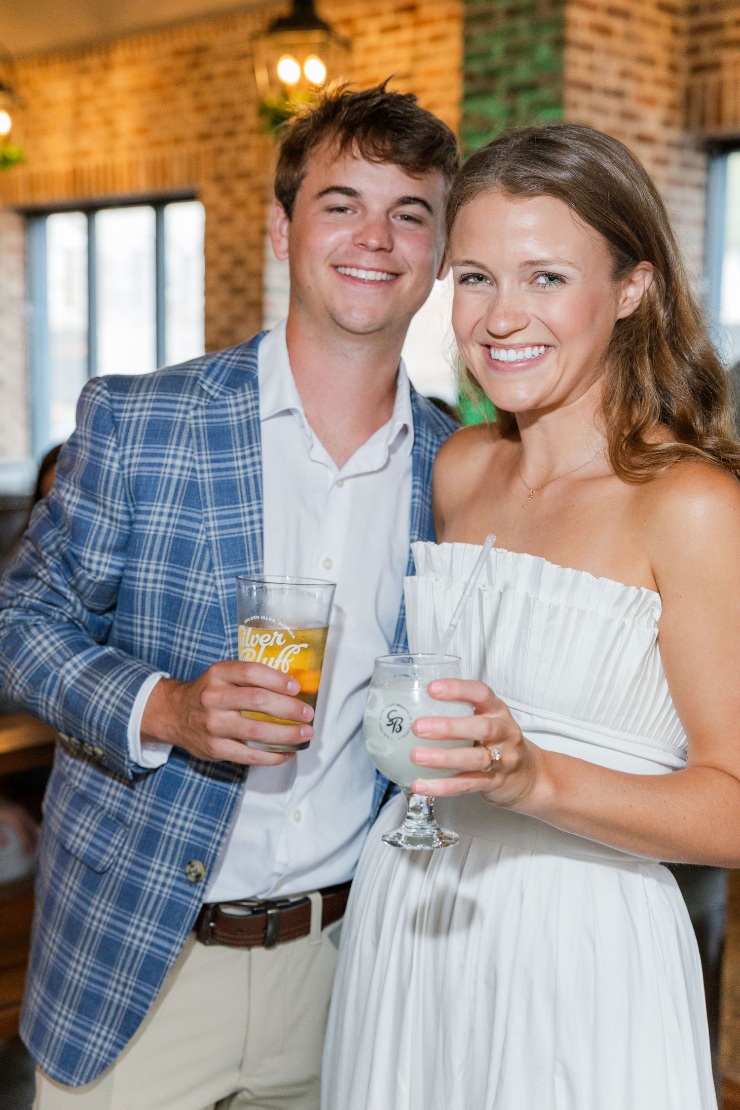 A smiling couple in formal attire holds cocktails at an indoor venue, man in blue plaid blazer, woman in white strapless dress.