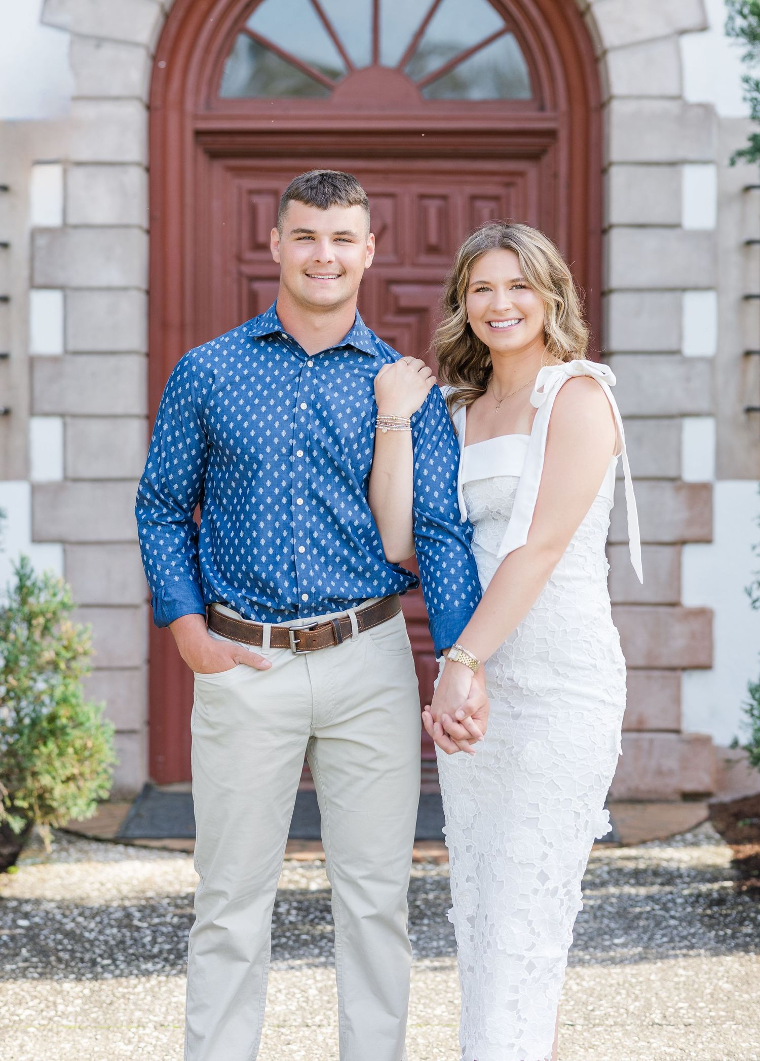 Smiling couple posing together in front of a red door, man in blue shirt and woman in white dress.