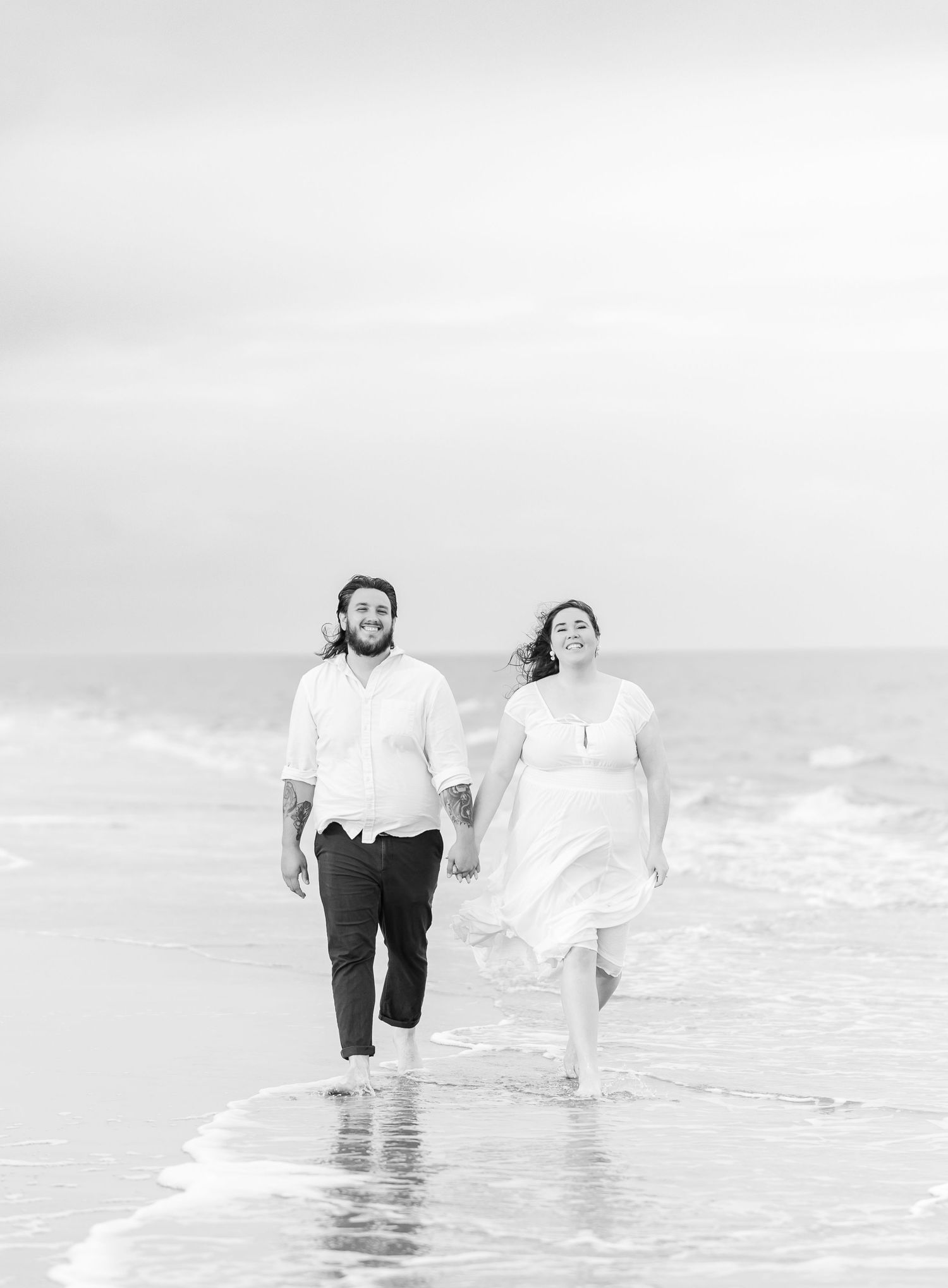 Happy couple walking through shallow ocean waves in black and white photography.