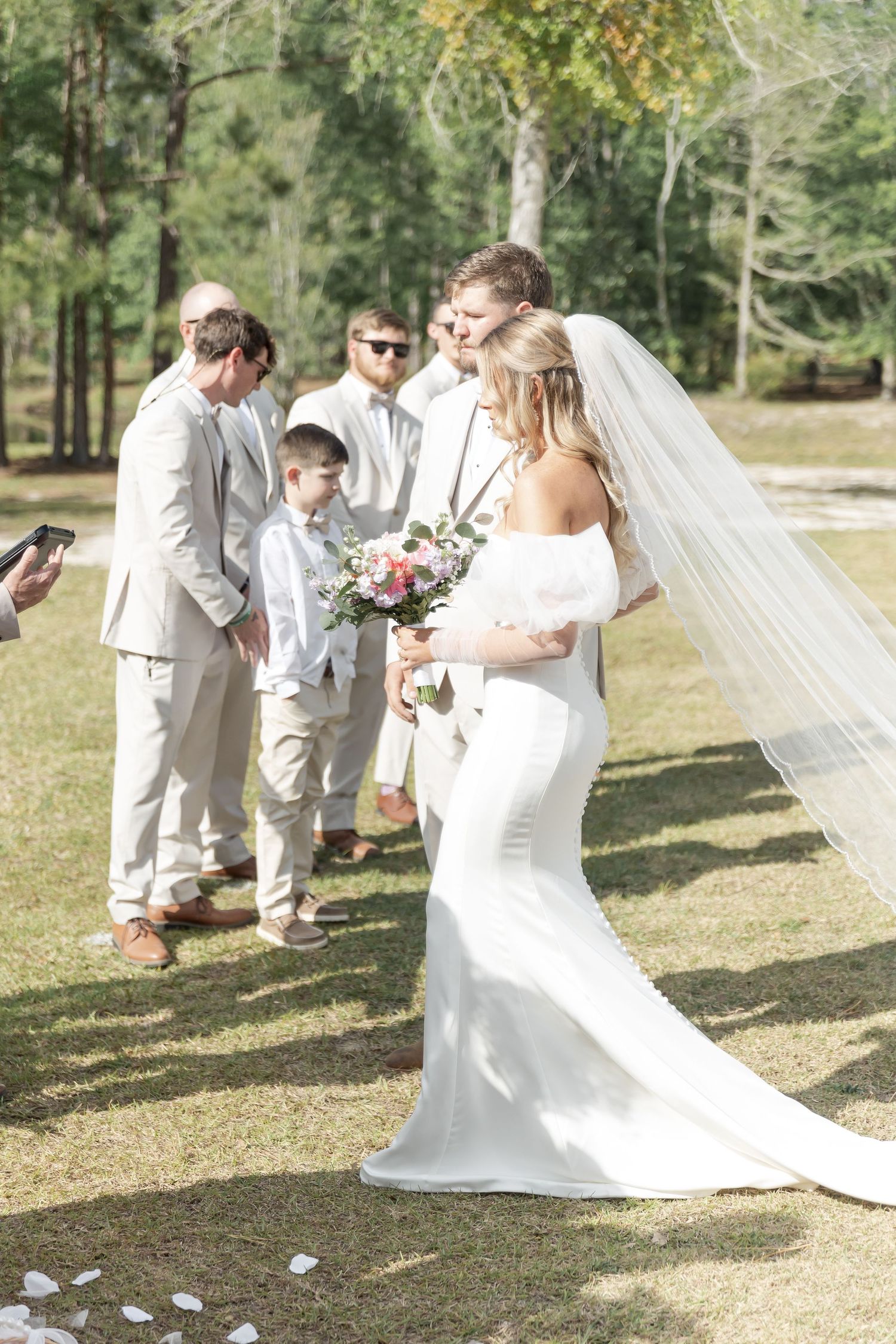 Wedding ceremony in progress with bridal party in light colored attire against wooded backdrop.