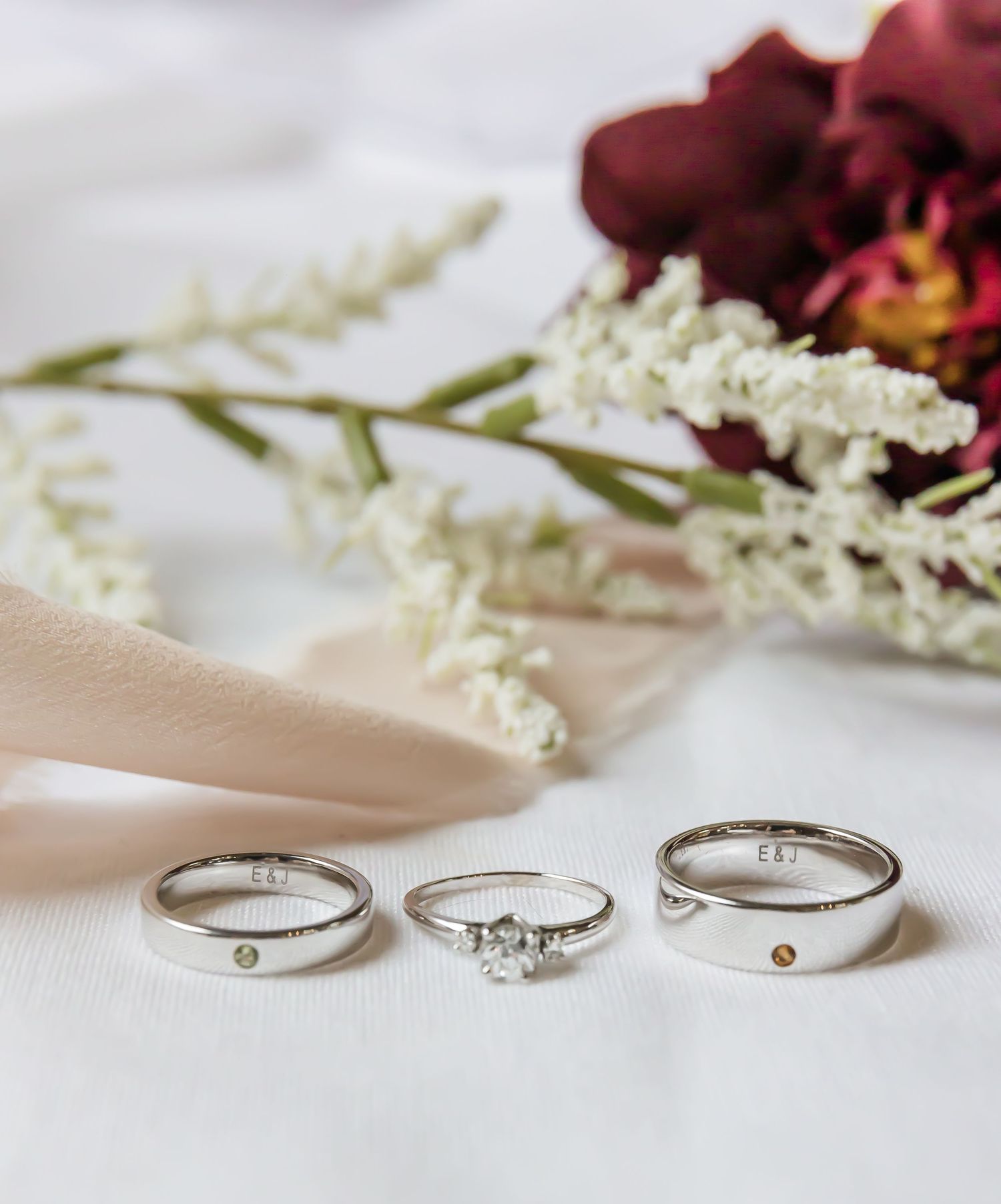 Three wedding rings displayed with burgundy and white flowers on white fabric.