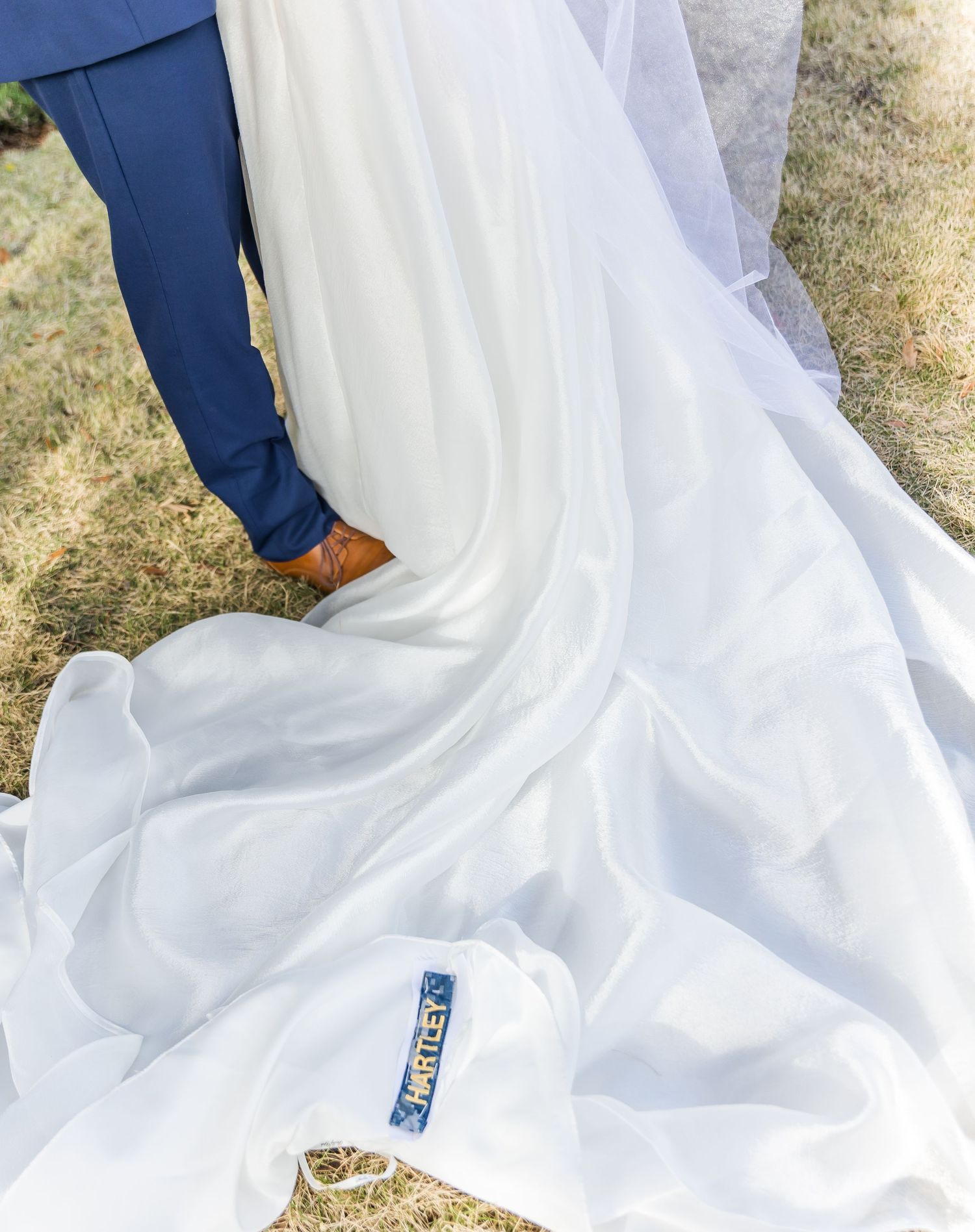 Close-up of flowing white wedding dress train against navy suit and brown shoes.