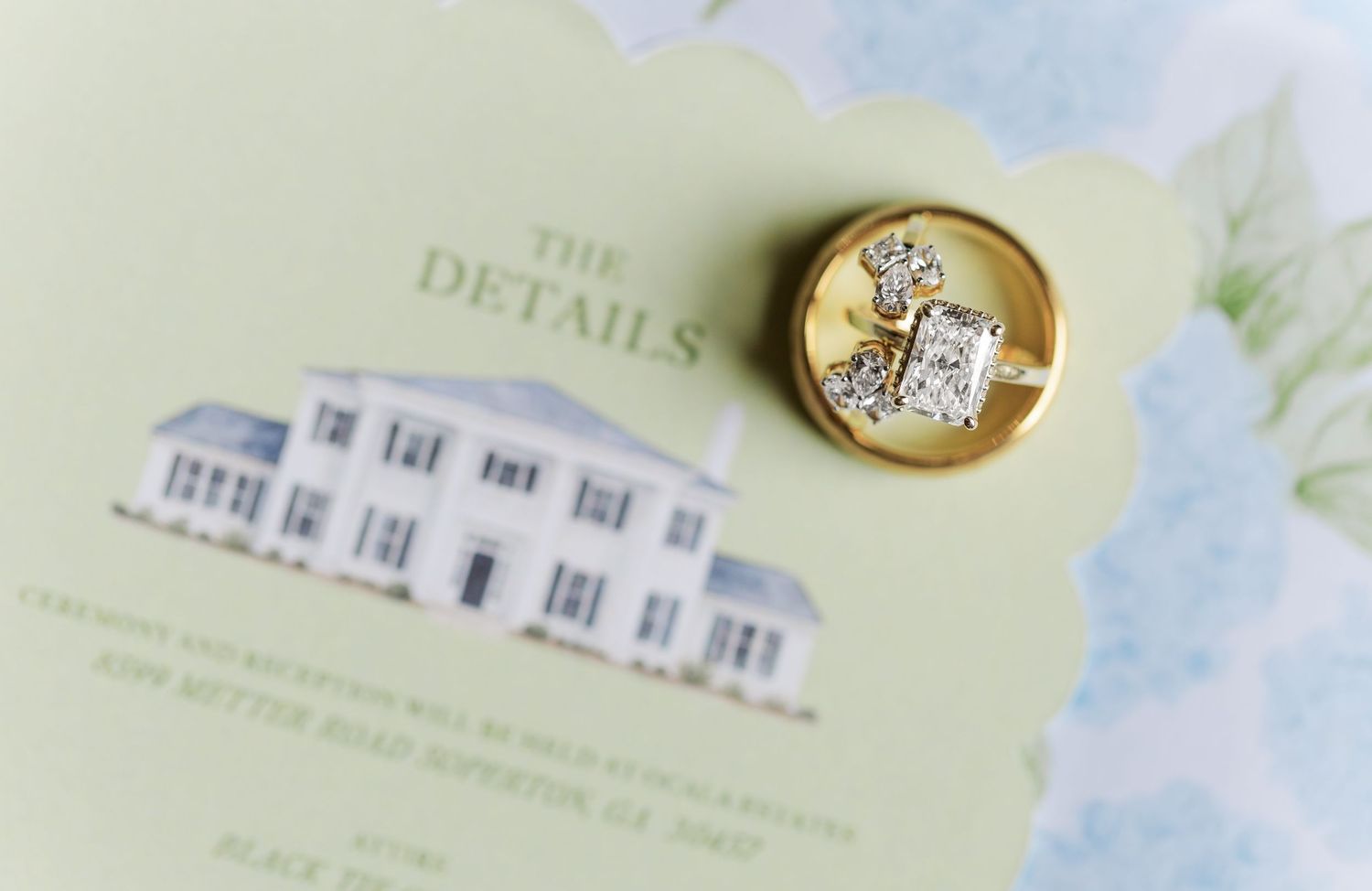 Gold diamond engagement ring resting on elegant wedding details card with floral design and venue illustration.
