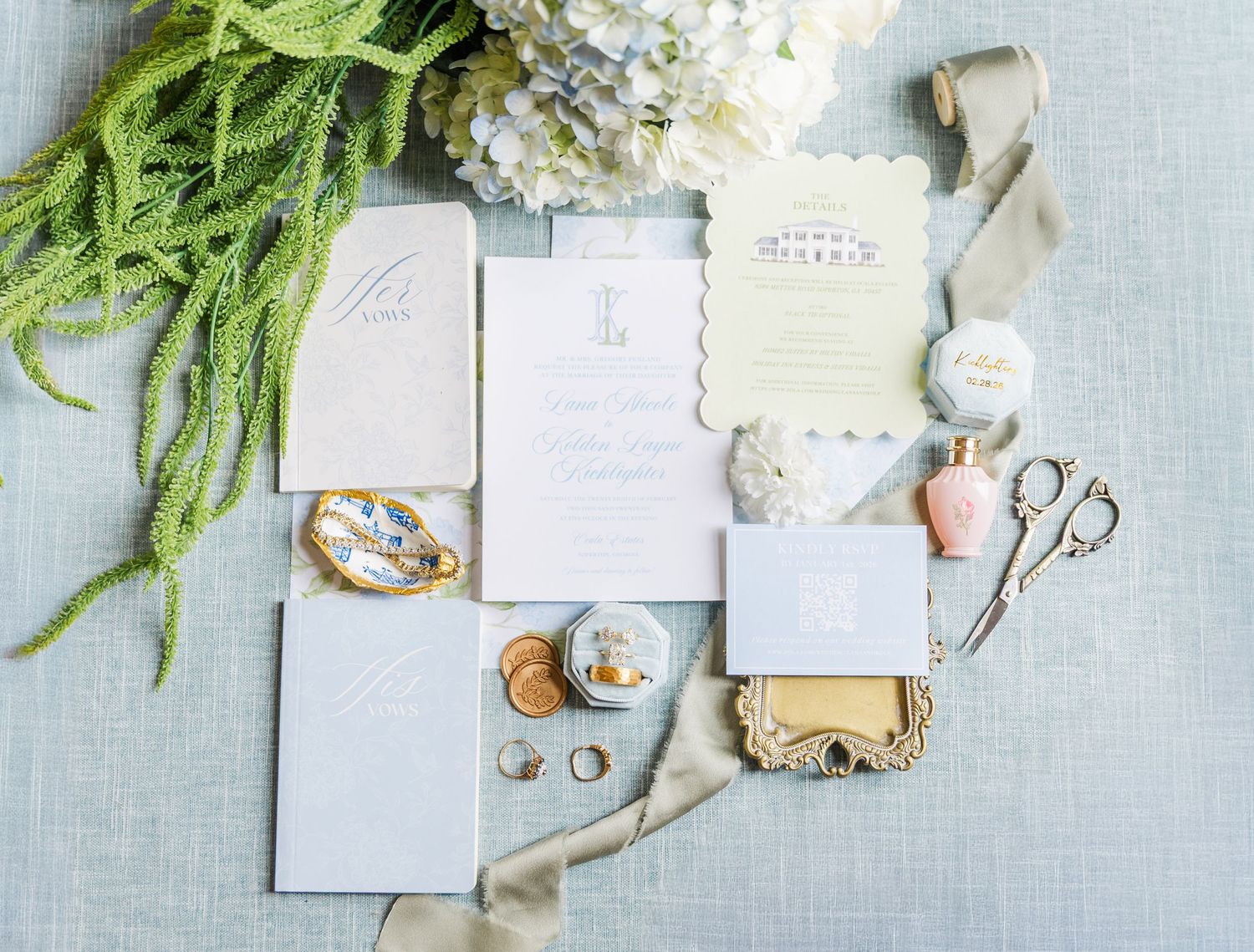 Elegant wedding invitation suite with dusty blue stationery, gold accents, scissors, rings, and greenery on a linen background.