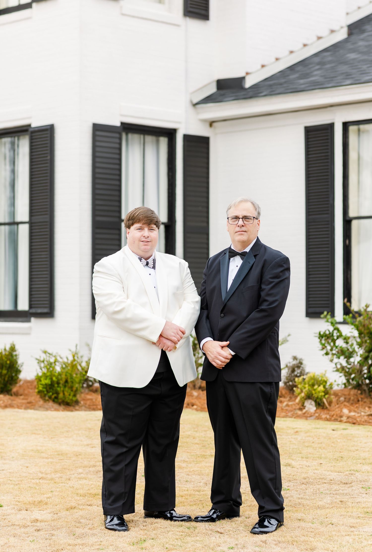 Groom in white tuxedo jacket stands with older man in black tuxedo outside white building with black shutters.
