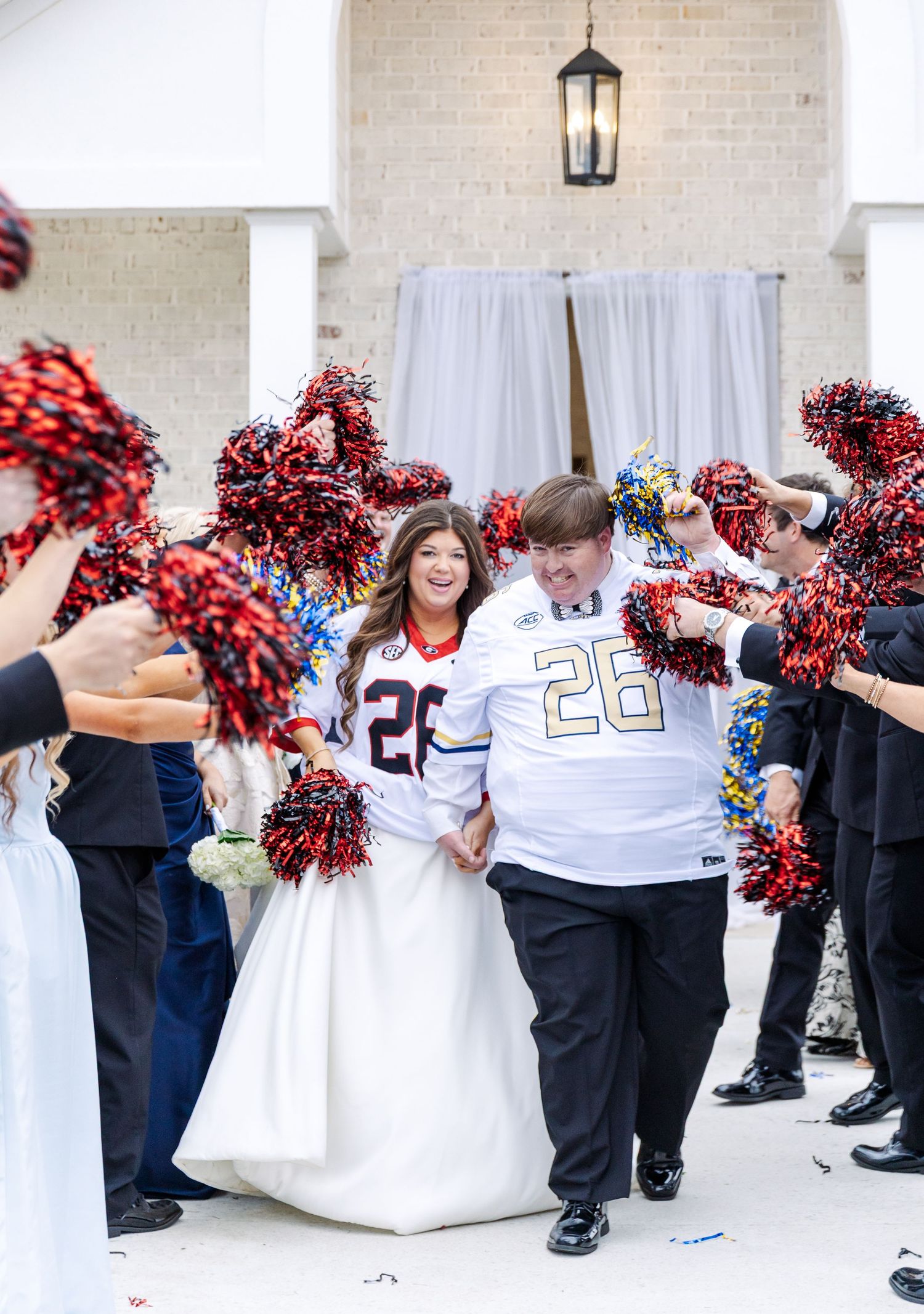 Newlyweds in football jerseys exit reception through tunnel of colorful pom-poms and streamers.